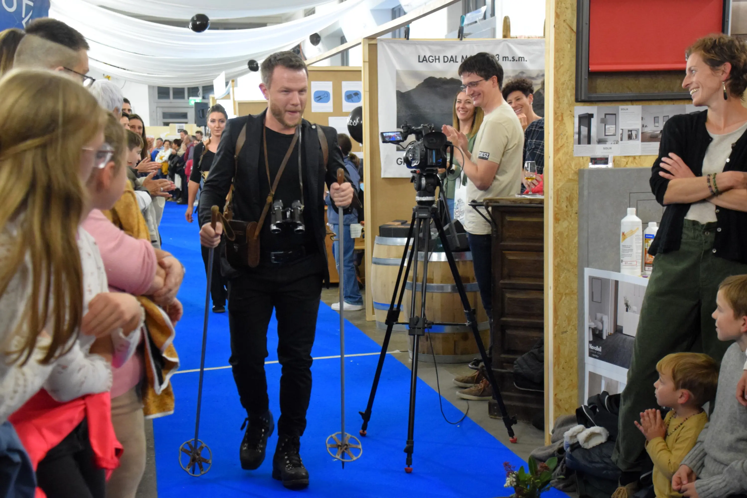 Un uomo cammina su una passerella blu, indossando una cintura con macchine fotografiche, mentre un gruppo di persone applaude. Sullo sfondo, un set di attrezzature da ripresa e una folla di spettatori.