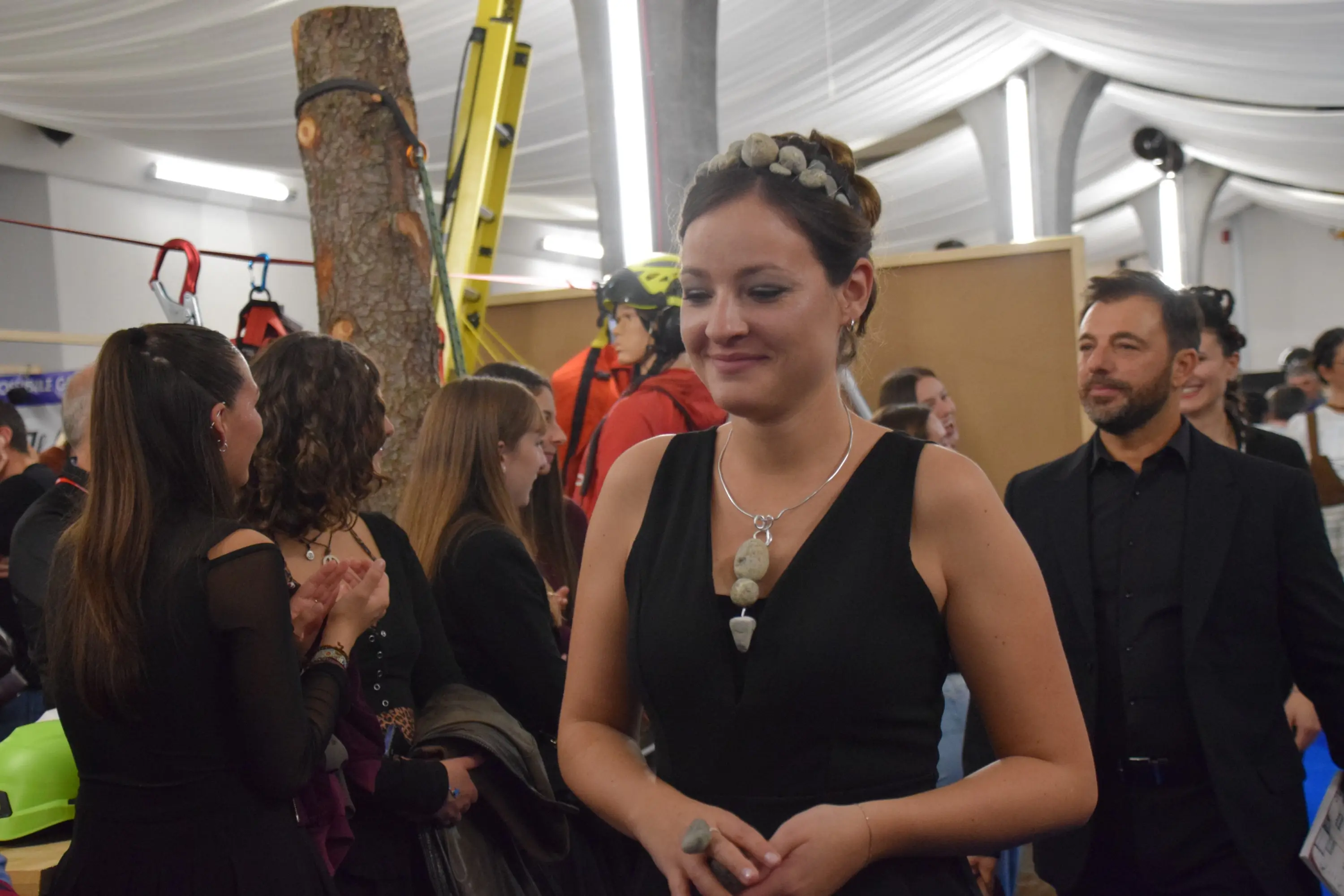 Una giovane donna in abito nero sorride, indossando gioielli particolari. Sullo sfondo, un gruppo di persone è impegnato in conversazioni, mentre alcune rovine di attrezzature per attività all'aperto sono visibili.