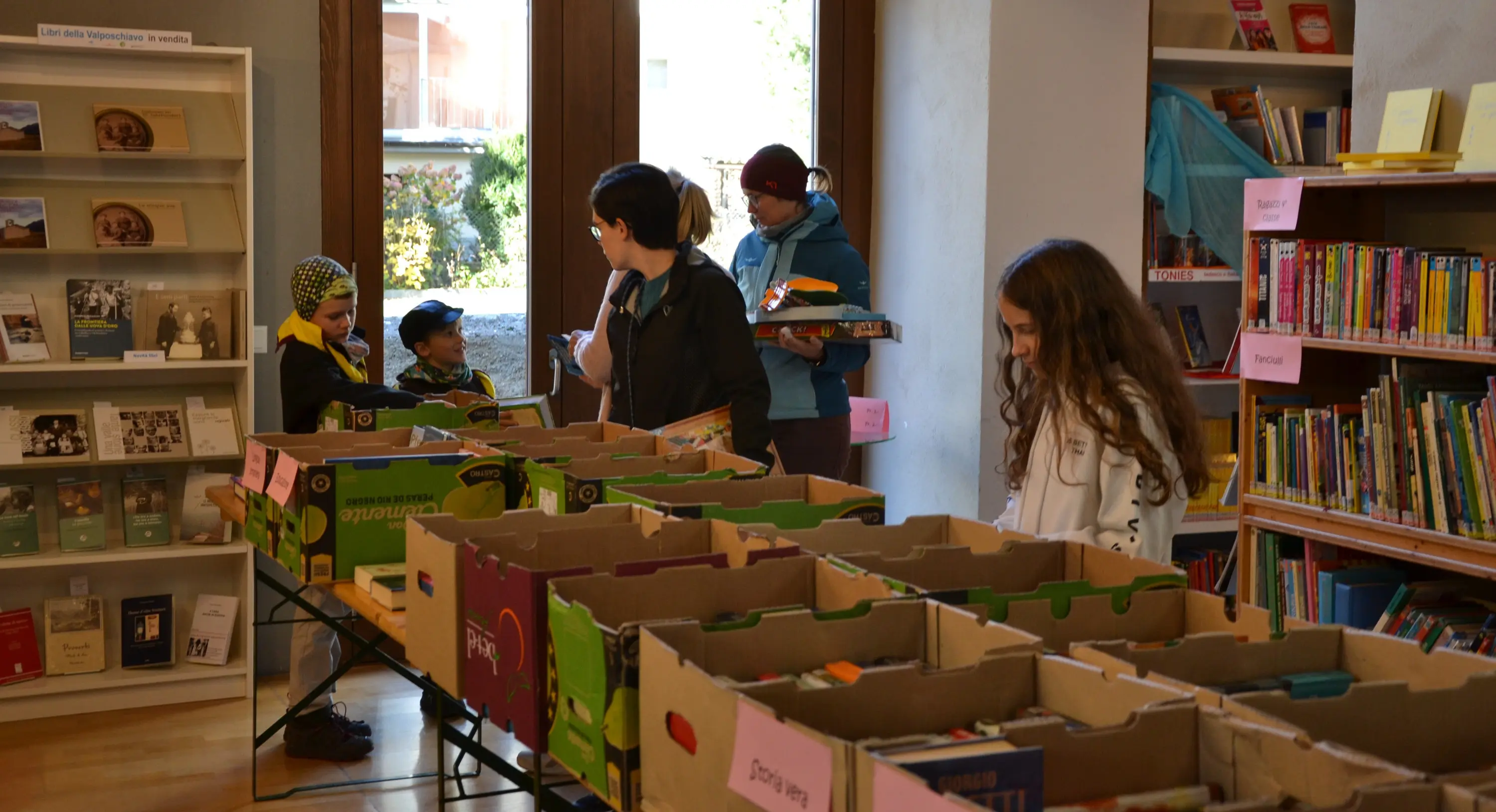 Una biblioteca con tavoli carichi di scatole di libri. Alcuni bambini e adulti sfogliano i libri, mentre uno dei bambini tiene un libro in mano. Sullo sfondo, scaffali pieni di altri libri.