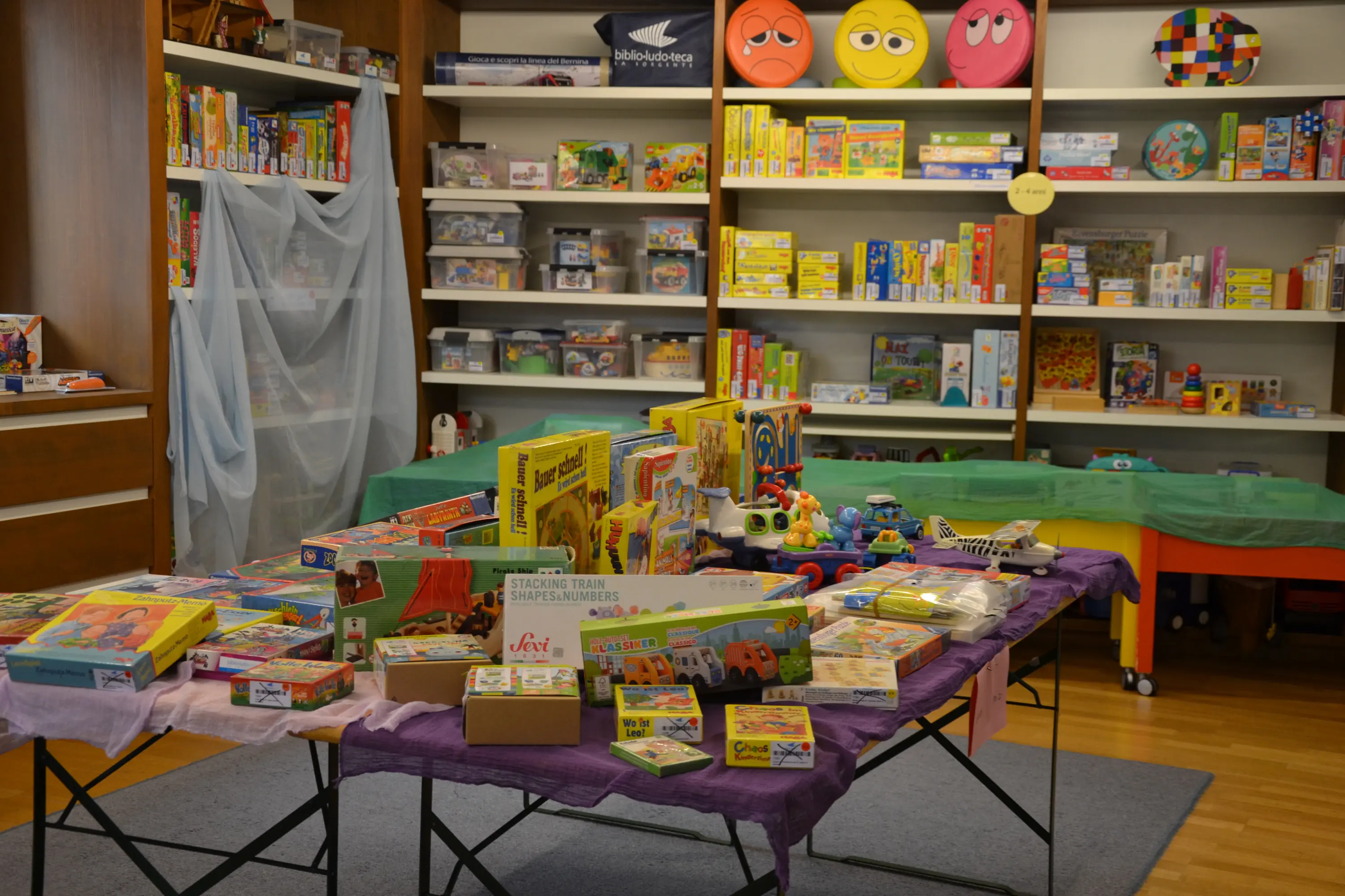 Una stanza ben illuminata con scaffali che contengono giochi e puzzle colorati. Un tavolo centrale è coperto di scatole di giochi e giocattoli vari, includendo automobili, puzzle e giochi didattici.