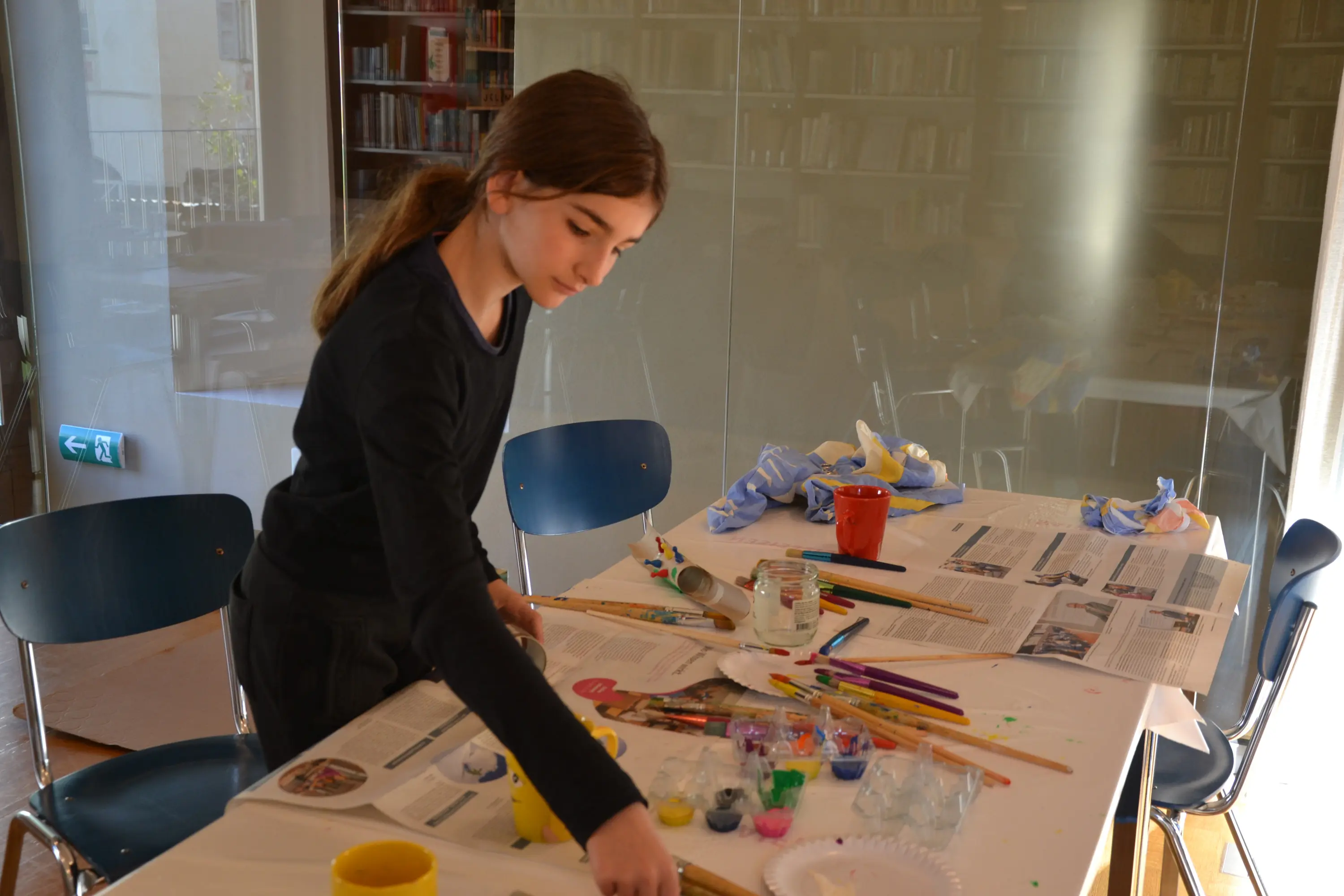 Una ragazza è intenta a dipingere su un tavolo ricoperto di materiali artistici, tra cui colori e pennelli. Sullo sfondo si intravedono scaffali con libri.