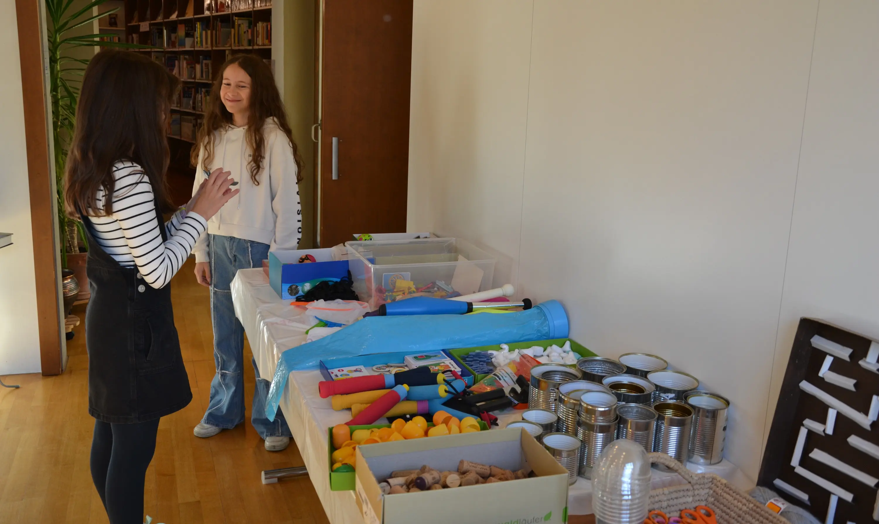 Due ragazze sorridenti conversano accanto a un tavolo carico di materiali per attività creative, tra cui scatole, colori, e oggetti vari. Sullo sfondo si intravede una libreria.