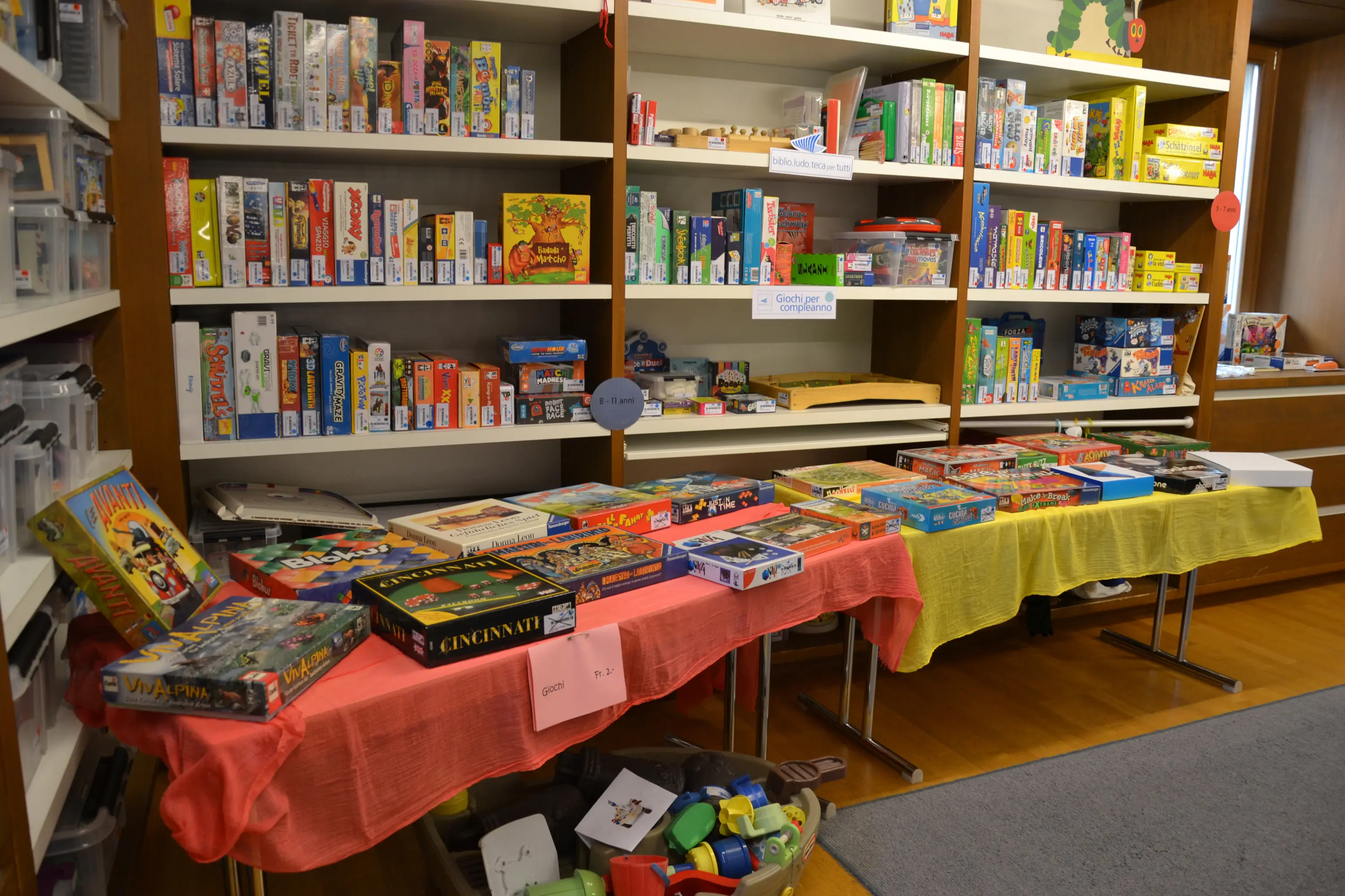 Una vista di una libreria con diverse scaffalature piene di giochi da tavolo e puzzle. Alcuni giochi sono esposti su tavoli coperti da tovaglie gialle e rosse.