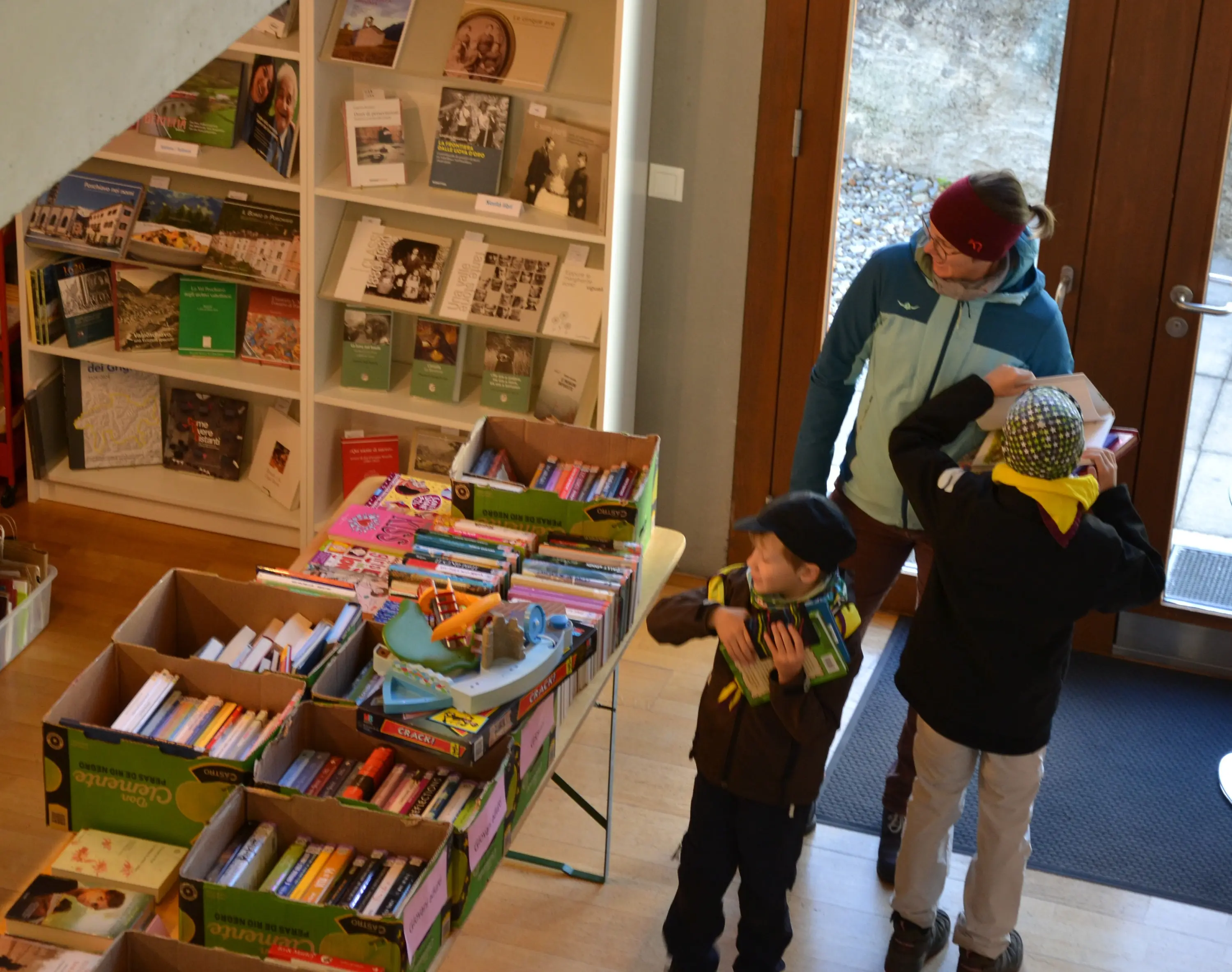 Due bambini e un adulto all'interno di una biblioteca, intenti a scegliere libri da scatole. Una delle scatole è piena di libri colorati, mentre l'adulto osserva i bambini. Sullo sfondo, scaffali con vari libri e materiali esposti.