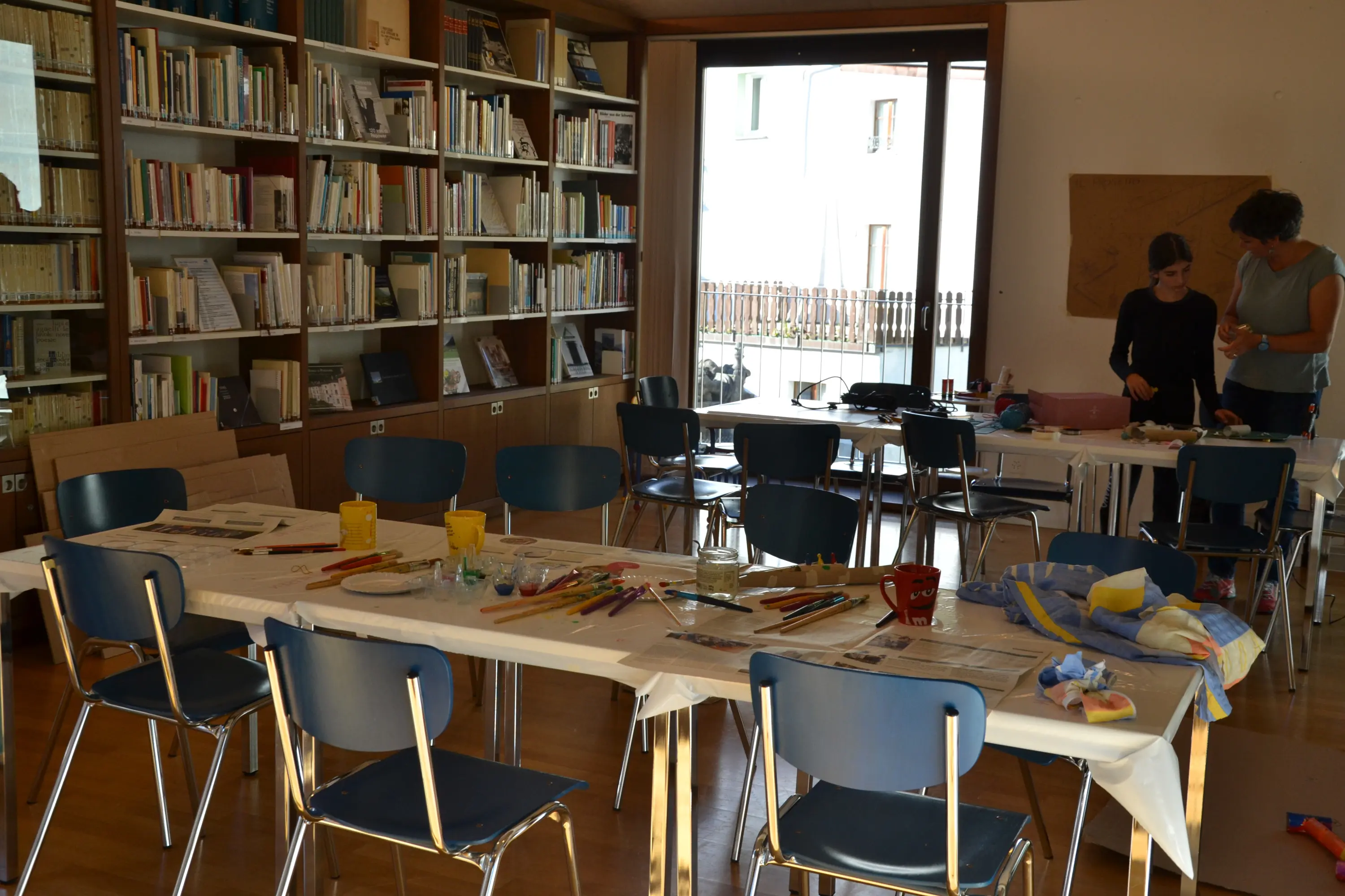 Una sala creativa con tavoli disposti per un laboratorio, circondati da scaffali di libri. Ci sono materiali artistici sparsi sui tavoli e due persone che lavorano insieme. La luce naturale entra dalle finestre.