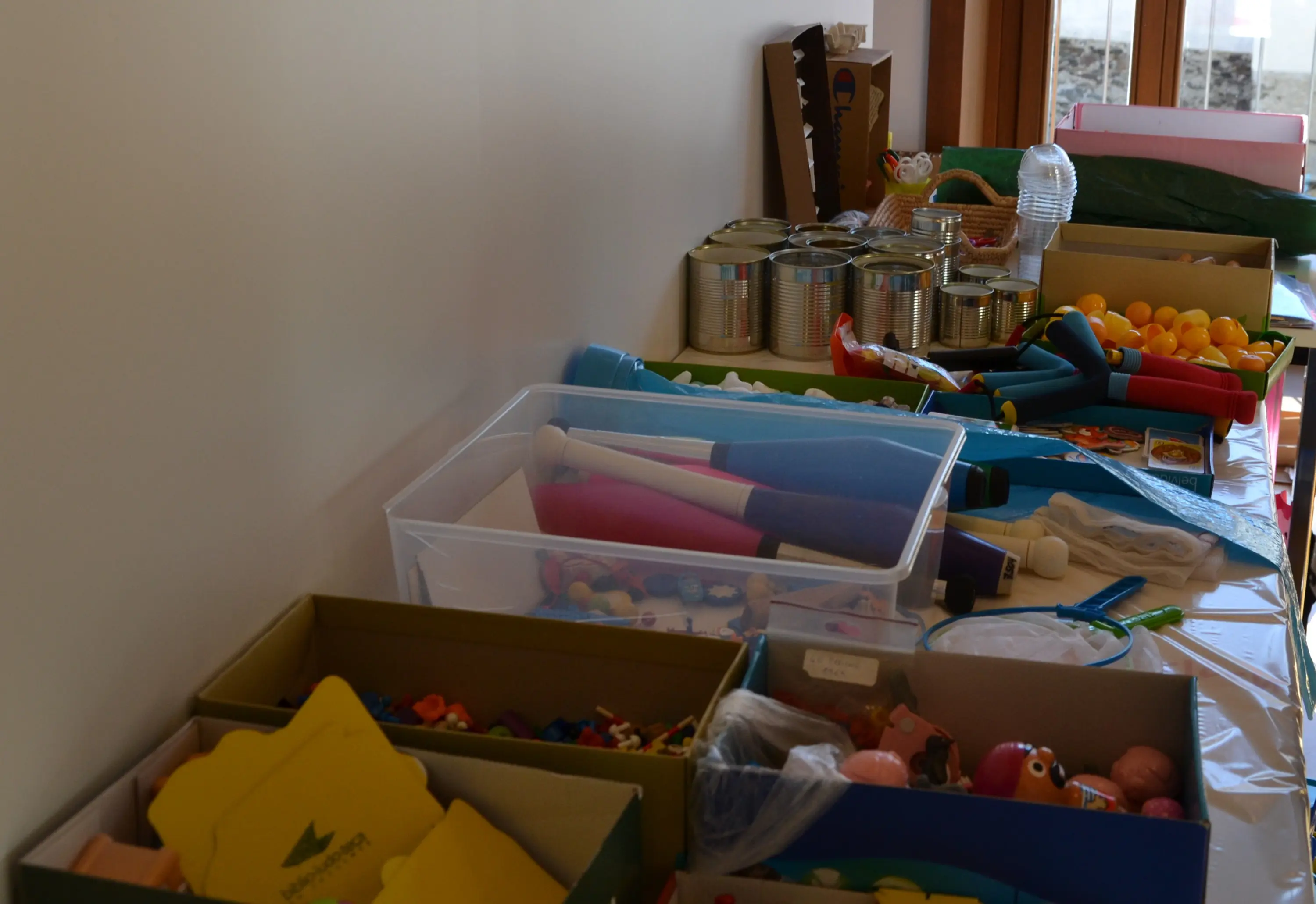 Variopinti materiali per attività manuali e giochi sono disposti su un tavolo. Ci sono scatole, contenitori trasparenti e una varietà di oggetti come strumenti musicali, palline e materiali di cancelleria.