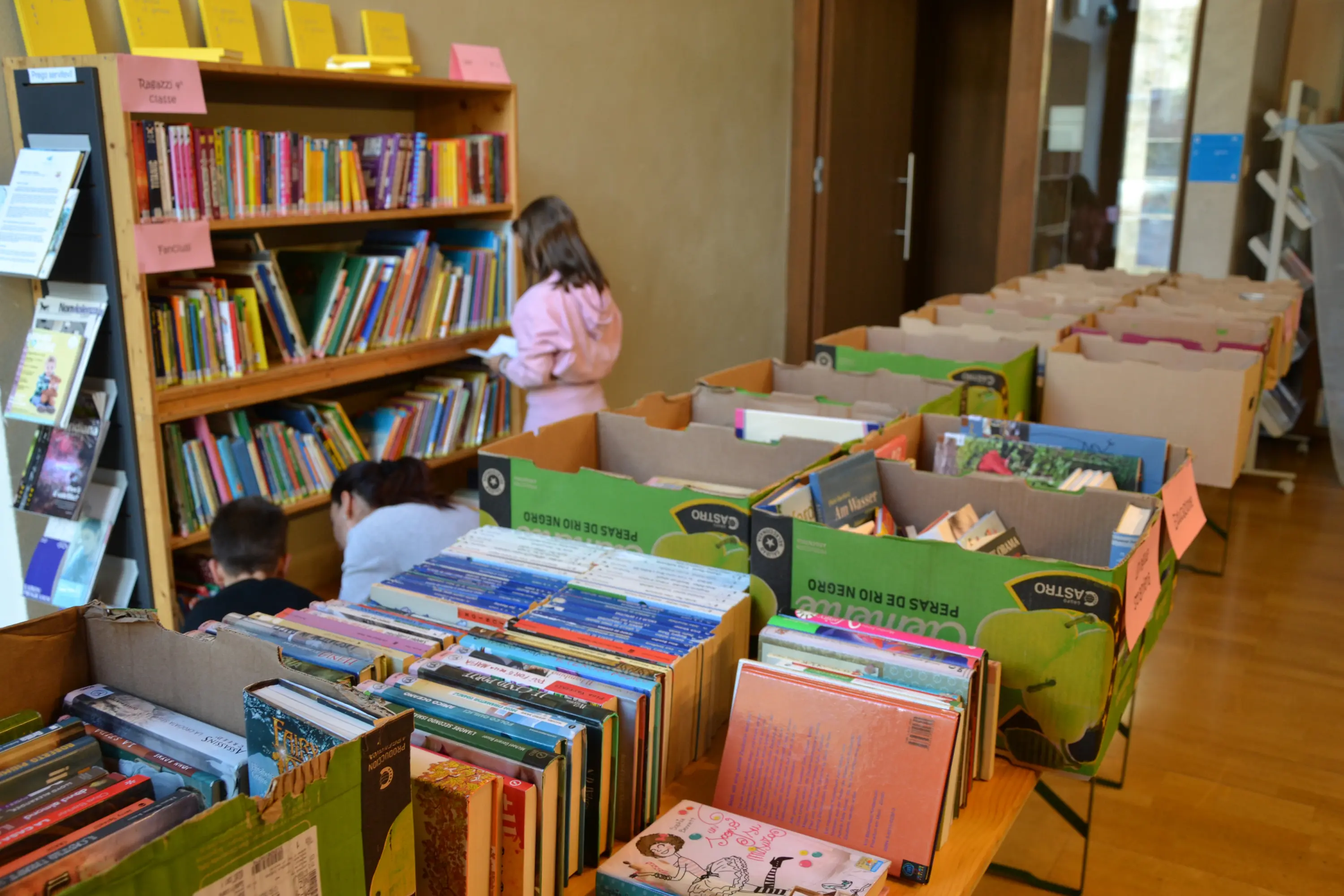 Una biblioteca con bambini che leggono. A sinistra, ci sono scatole di libri ordinati, mentre sullo sfondo si vede uno scaffale di libri con diverse copertine colorate.