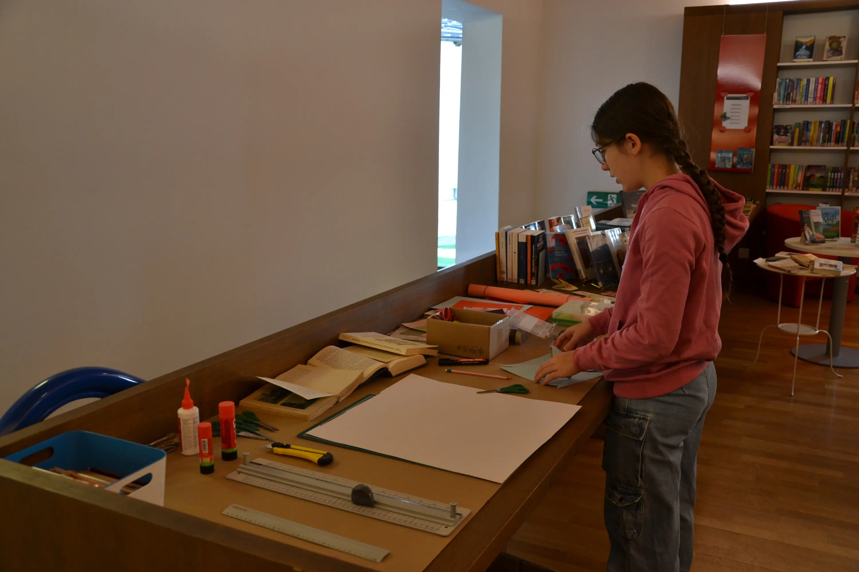 Una ragazza con occhiali sta lavorando su un tavolo di legno, circondata da materiali per il bricolage come colla, forbici e fogli di carta. Sullo sfondo ci sono scaffali pieni di libri.