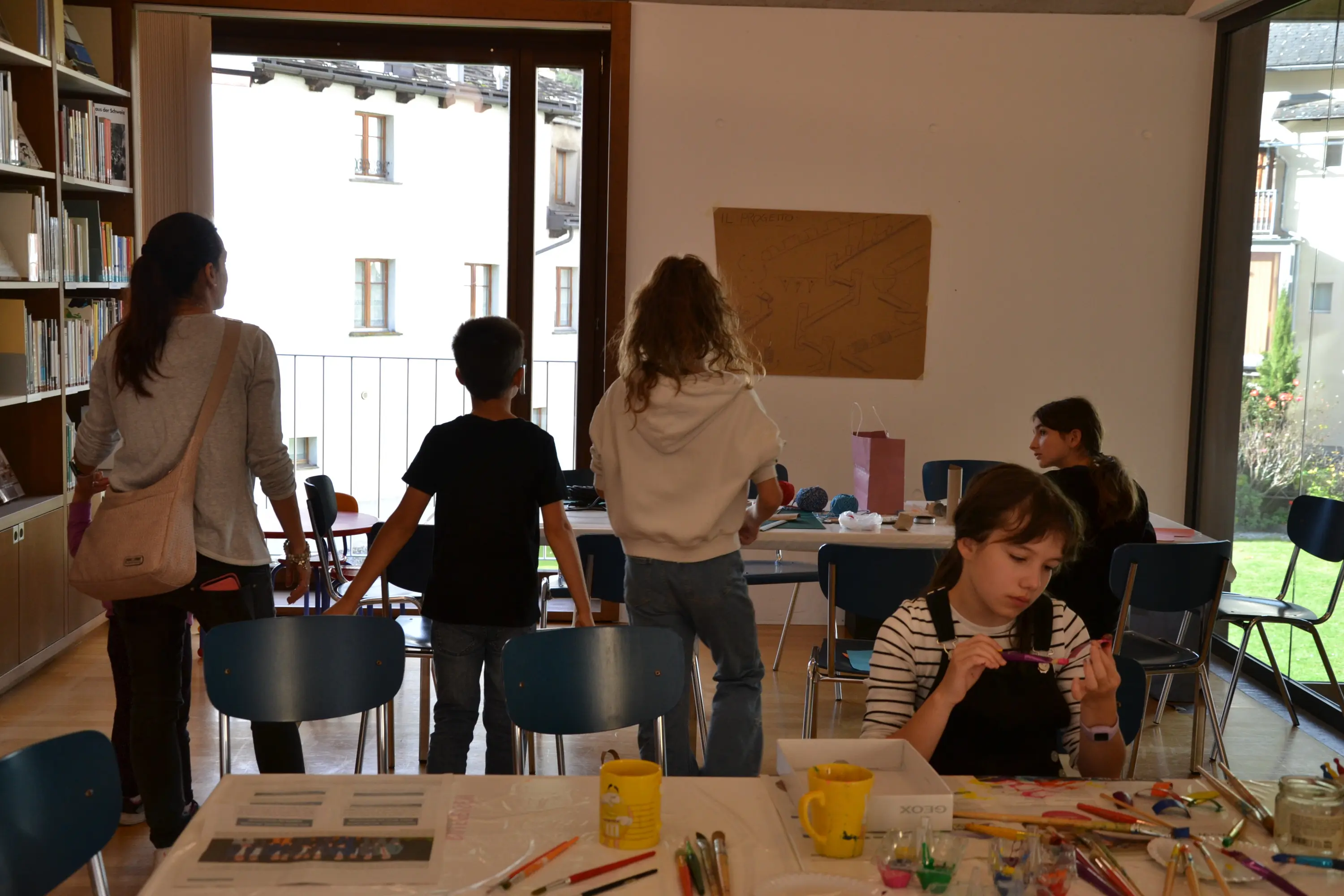 Due adulti e tre bambini in un'aula creativa. I bambini sono in piedi mentre una ragazza seduta dipinge su un tavolo. Una grande finestra offre una vista su un giardino.