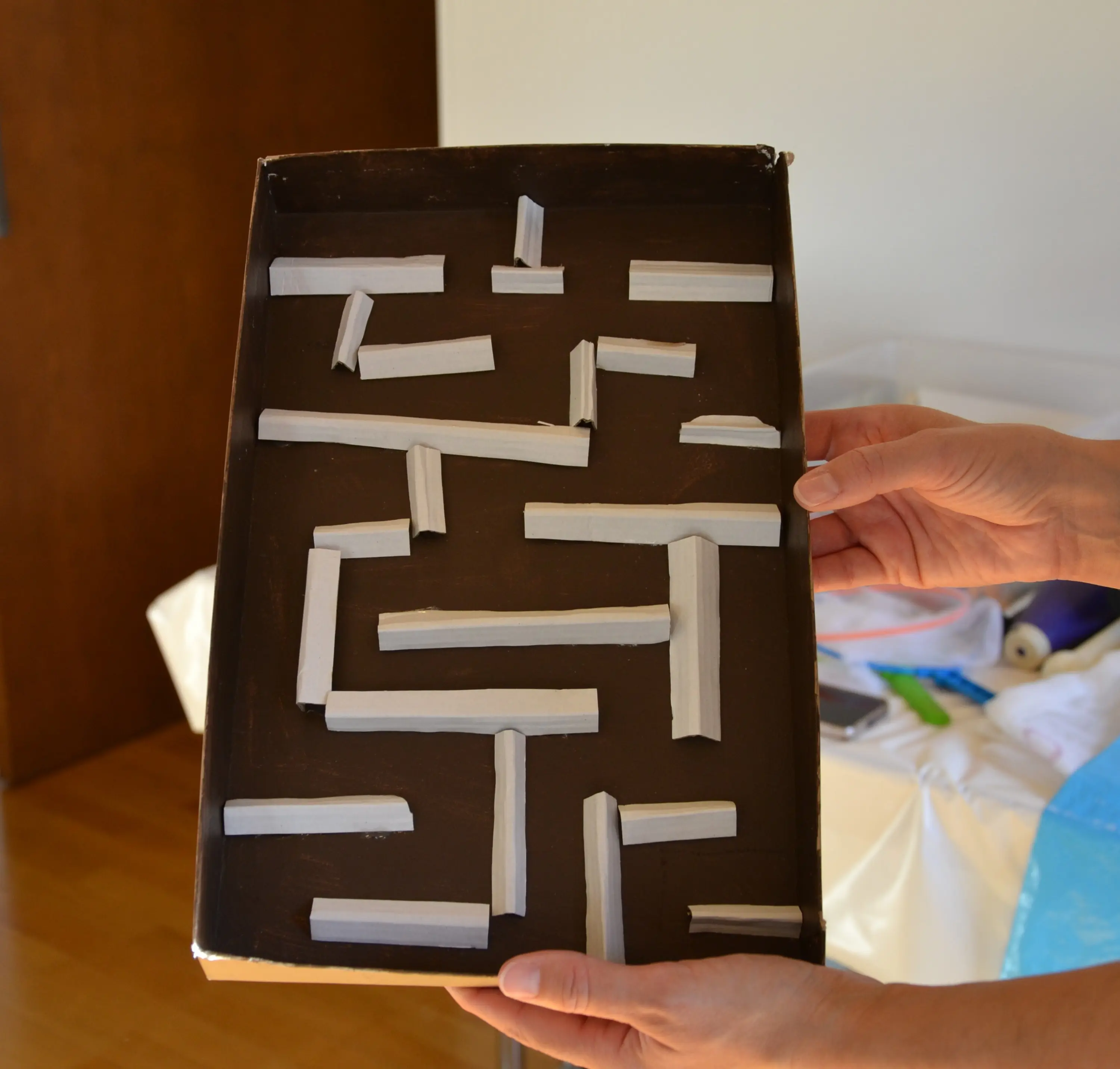 Un labirinto realizzato con strisce di nastro adesivo su una base scura, tenuto in mano da una persona. Si possono vedere vari percorsi e ostacoli.