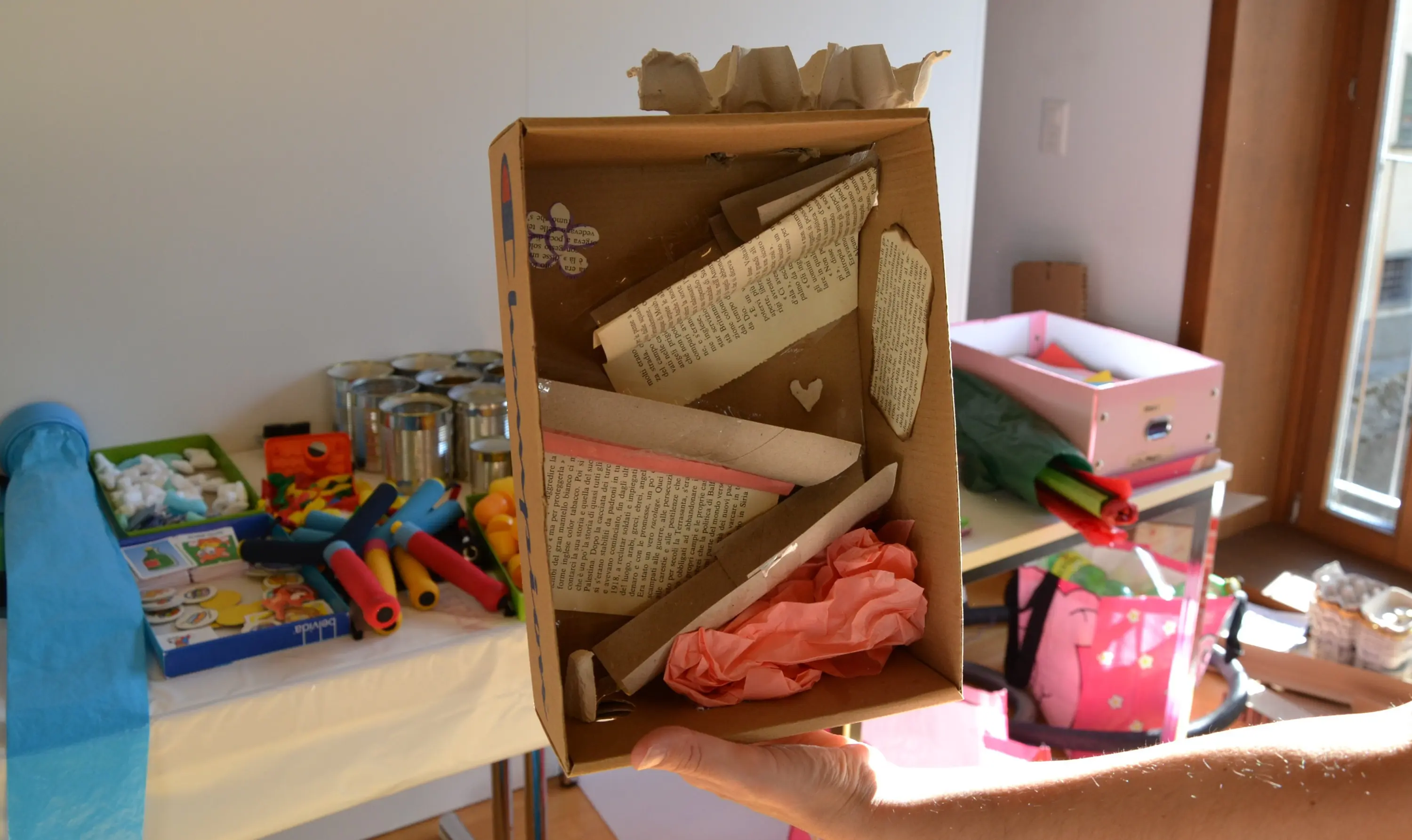 Una scatola di cartone contenente ritagli di carta, pagine di libri e carta velina, sorretta da una persona. Sullo sfondo si vedono materiali creativi come rotoli di carta e scatole colorate.
