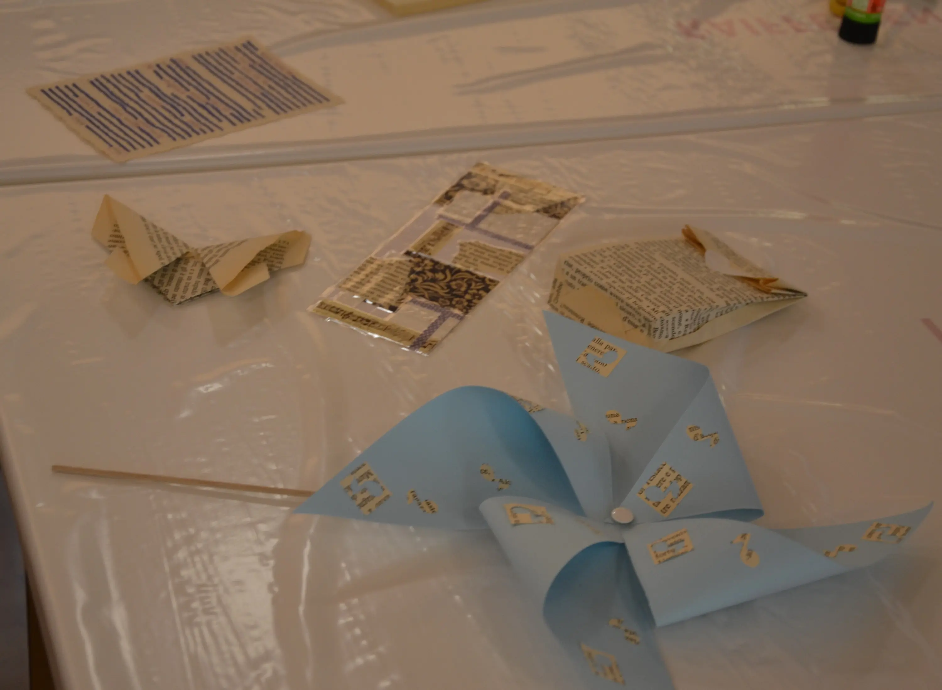 Vari oggetti artigianali su un tavolo. Ci sono forme piegate di carta, una girandola blu con simboli, e vari pezzi di carta decorativi.