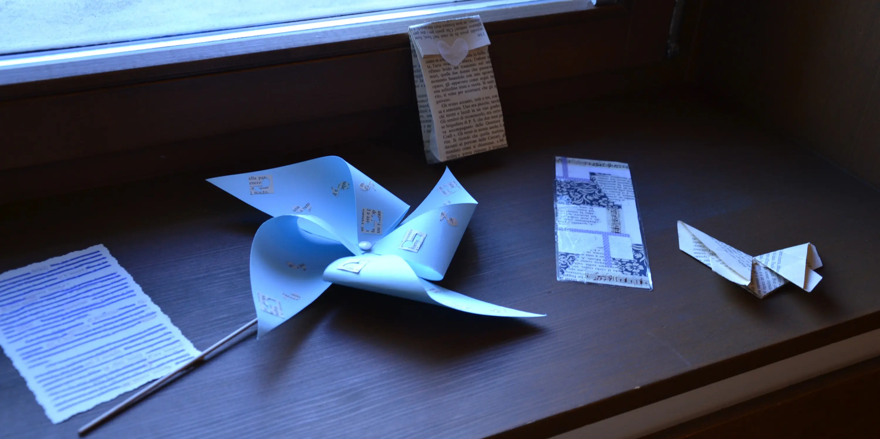 Variopinti origami e lettere in carta disposti su un davanzale. Un grande origami blu si distingue al centro, circondato da pezzi di carta stampata e piegata in forme diverse. La luce entra dalla finestra, illuminando i dettagli dei pezzi.
