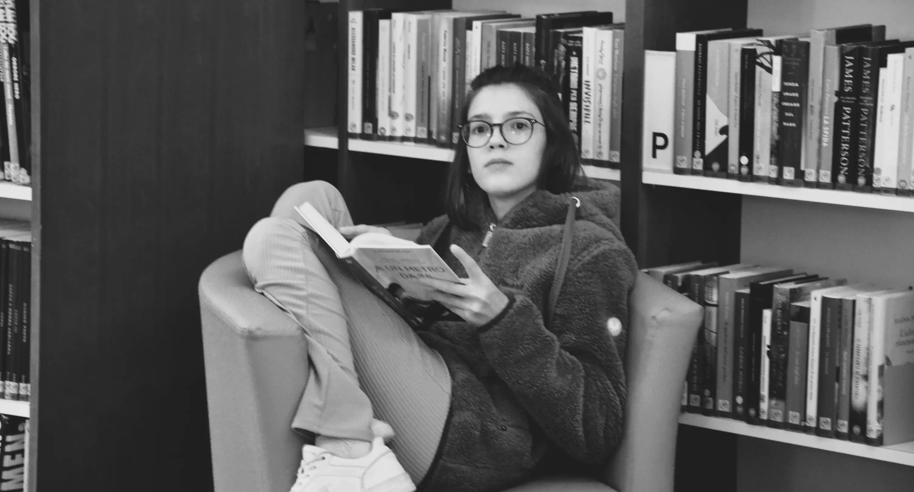 Ragazza seduta su una poltrona in biblioteca, che legge un libro con un'espressione concentrata. Scaffale di libri sullo sfondo.