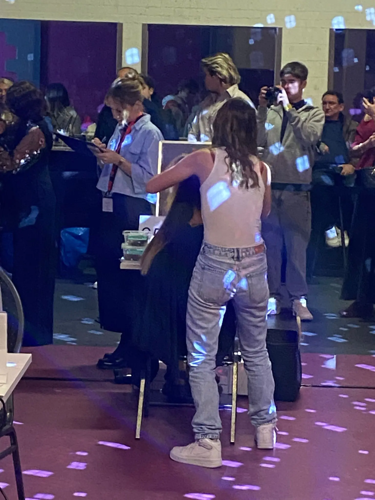 Un gruppo di persone in un ambiente vivace. Una donna con i capelli lunghi e ricci sta sistemando i capelli di un'altra persona. Sullo sfondo, altre persone sono coinvolte in attività varie, con luci colorate che creano un'atmosfera festosa.