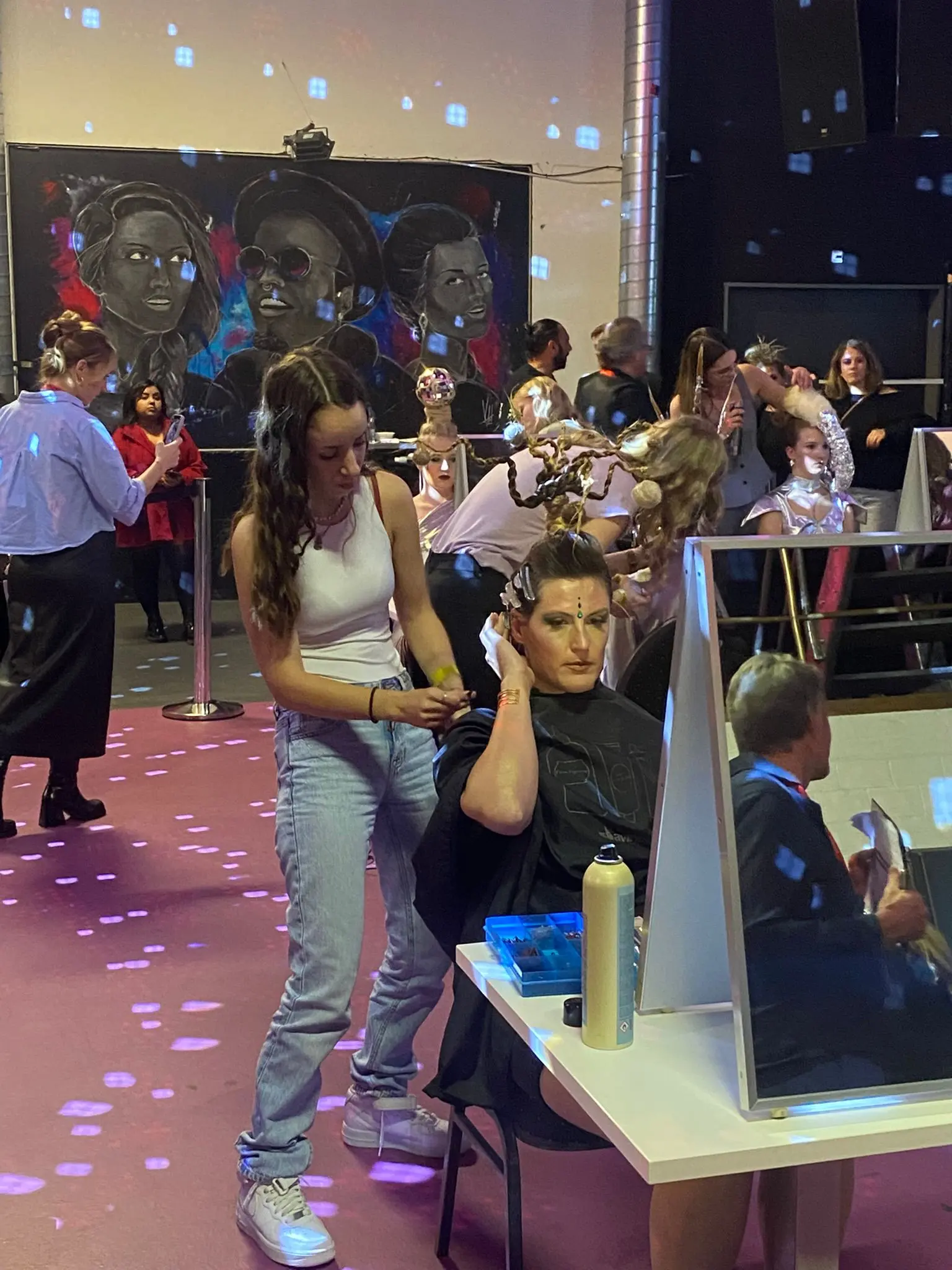 Una persona sta acconciando i capelli di un’altra mentre diverse persone sono impegnate in attività di bellezza in un ambiente vivace. Sullo sfondo, ci sono opere d'arte murale e luci colorate che creano un'atmosfera festiva.