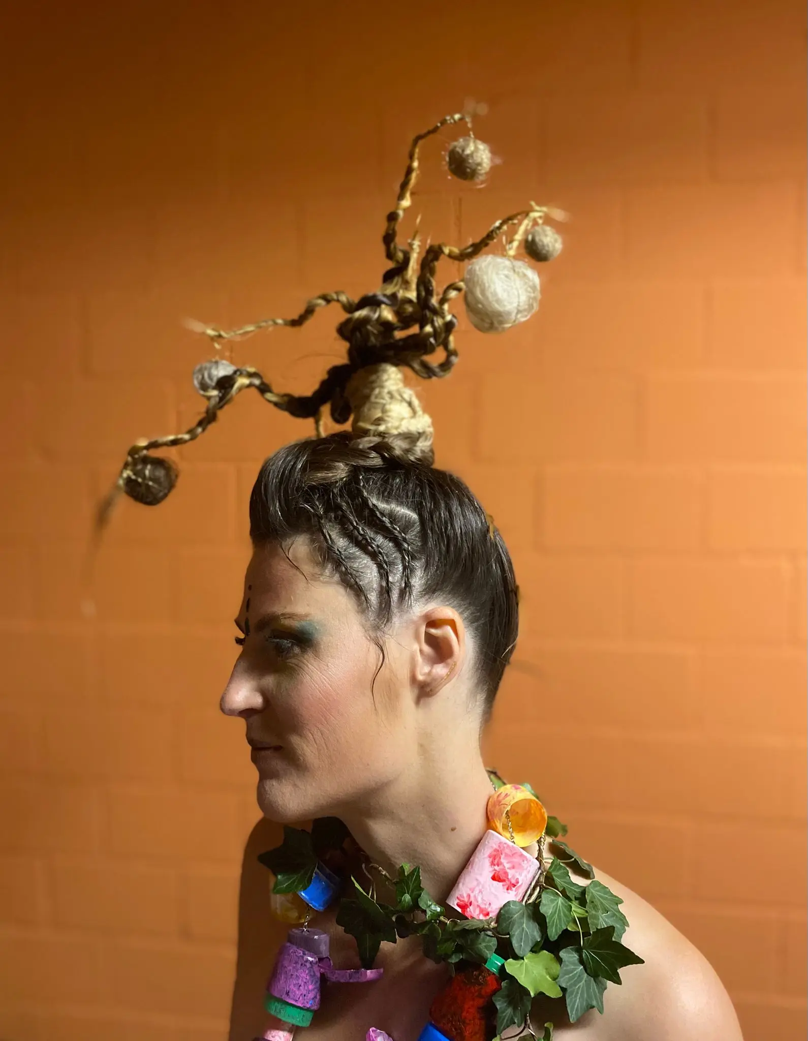Una donna con lunghi capelli raccolti in un acconciatura elaborata, adornata con elementi naturali come rami e palline di fibra. Indossa una collana colorata composta da materiali vari. Lo sfondo è arancione.