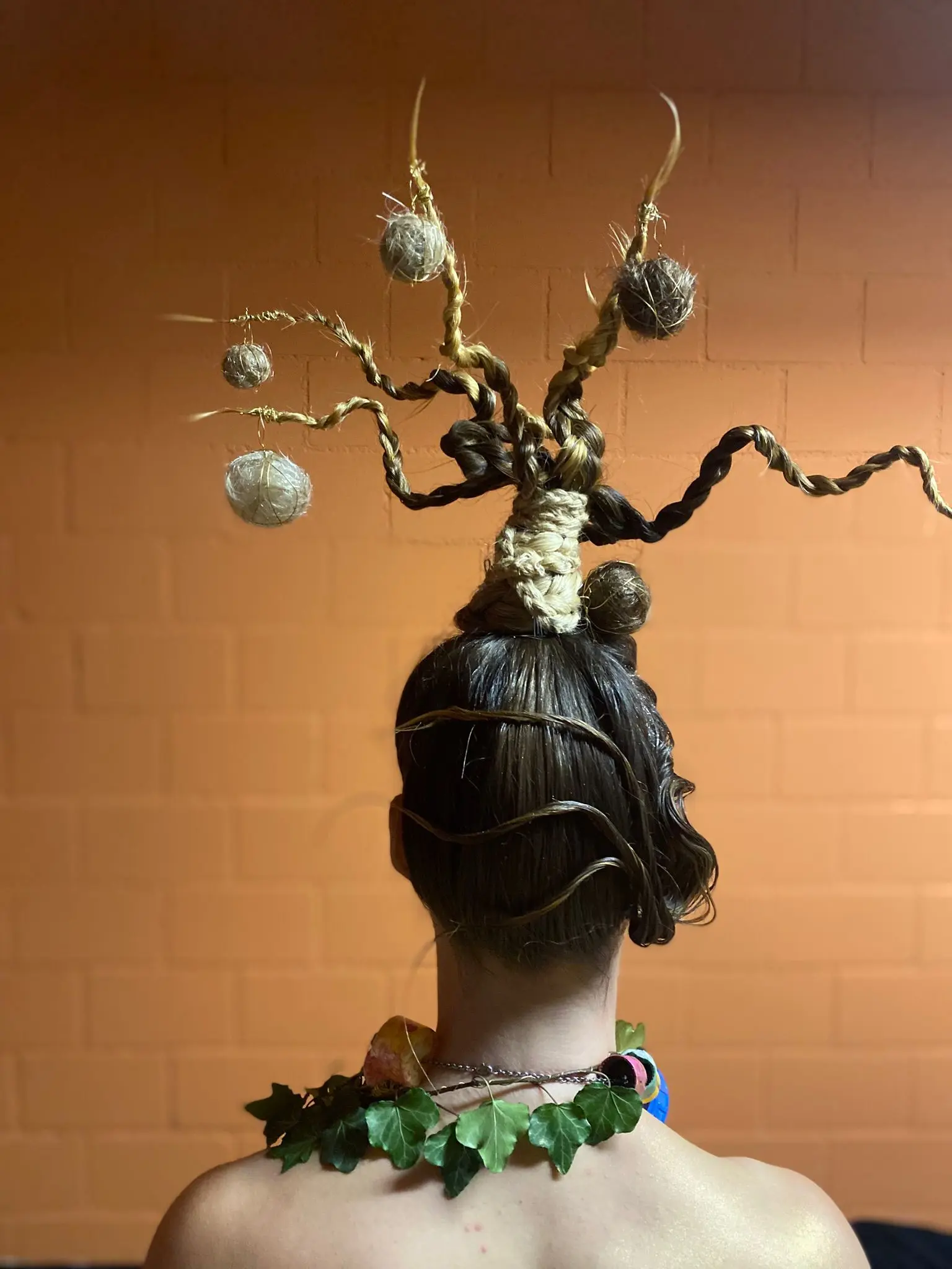 Acconciatura creativa con stile ispirato agli alberi, caratterizzata da ciocche intrecciate e decorazioni che ricordano noci. Collana di foglie verdi completa il look.