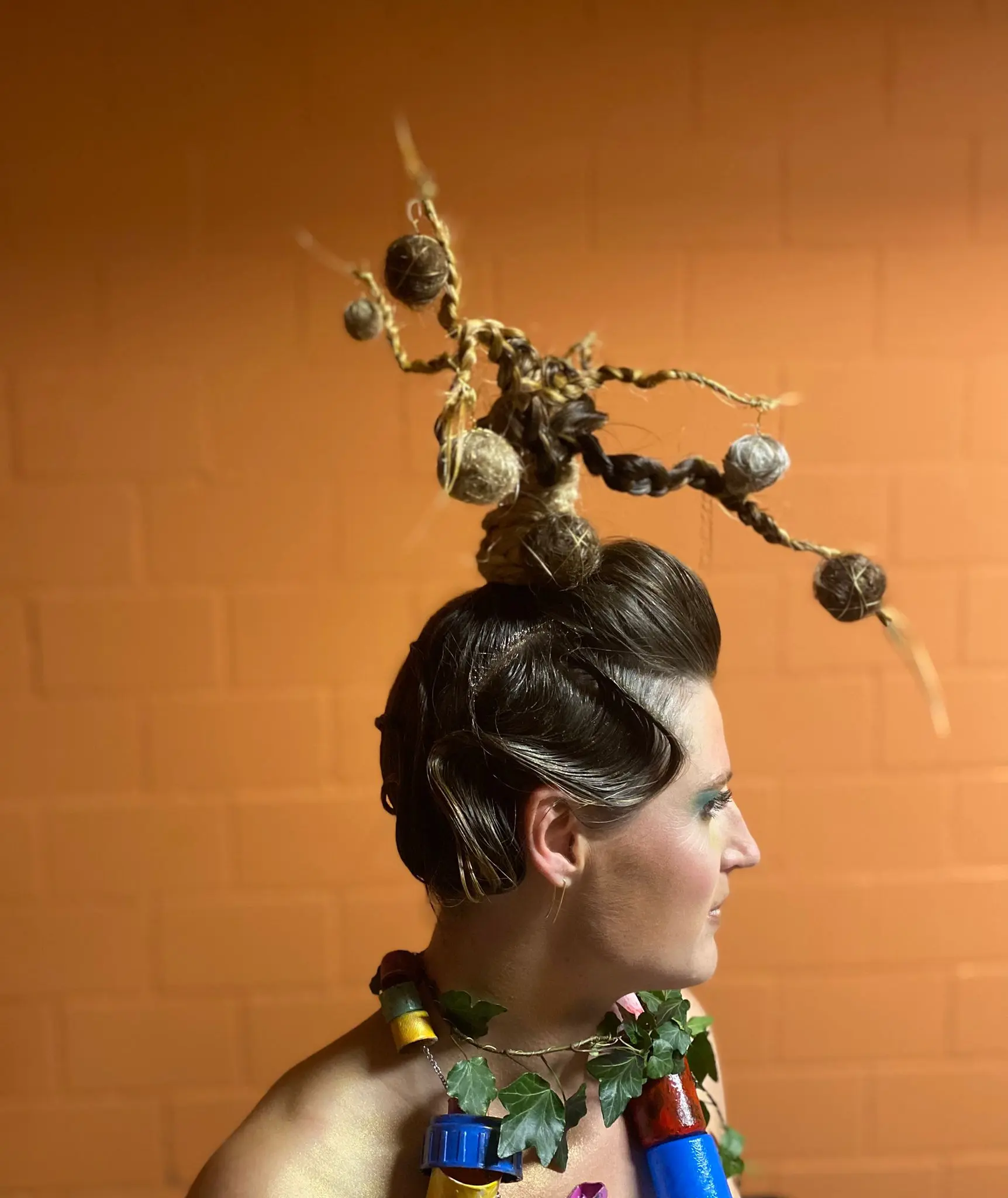 Acconciatura artistica con capelli raccolti e decorazioni naturali. Soggetto indossa un collier di materiali colorati e foglie. Sullo sfondo, una parete arancione.