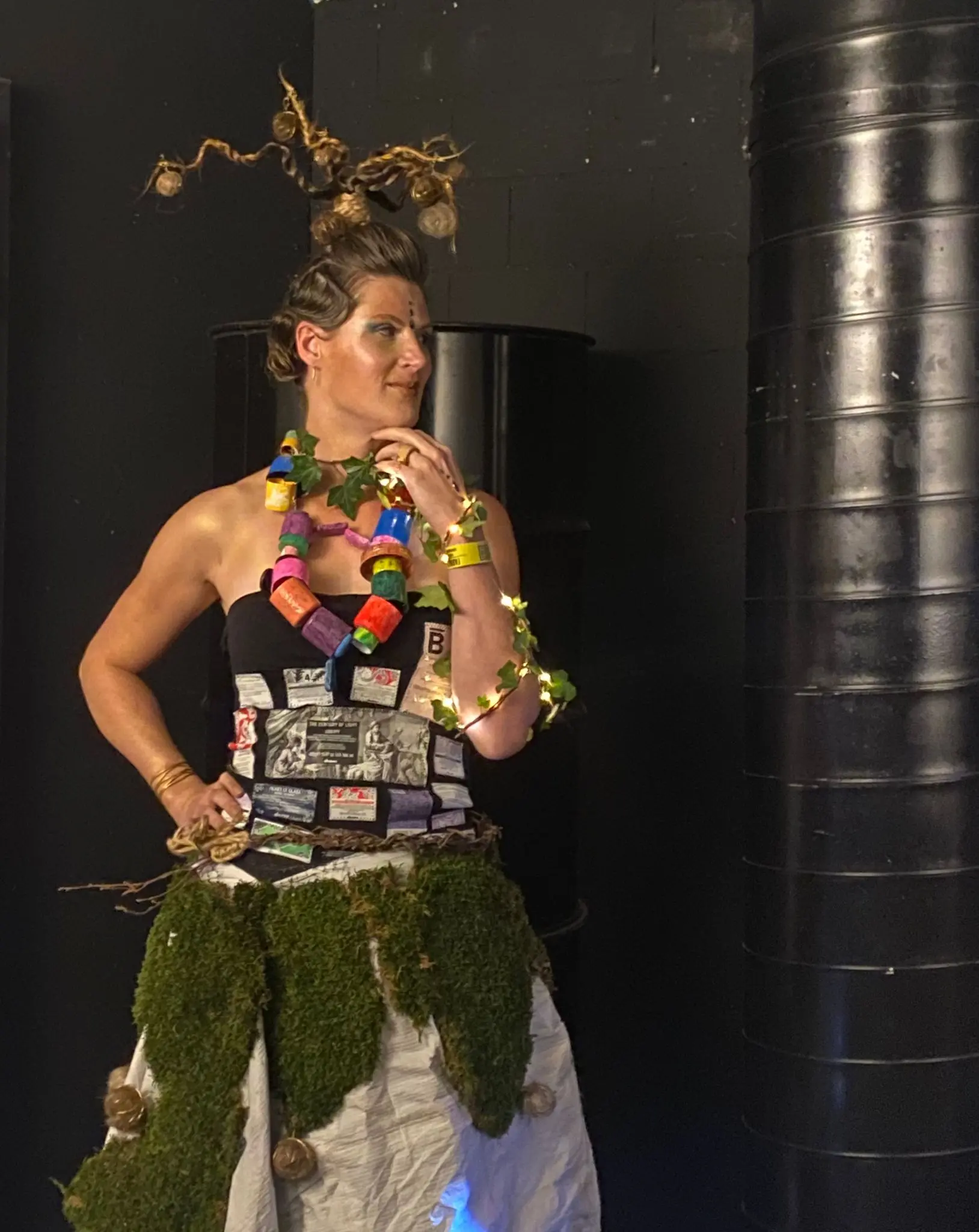 Donna in piedi con un abito creativo decorato con muschio e oggetti vari. Indossa un collier di colori vivaci e ha una acconciatura con rami e decorazioni luminose. Sullo sfondo, ci sono elementi industriali scuri.