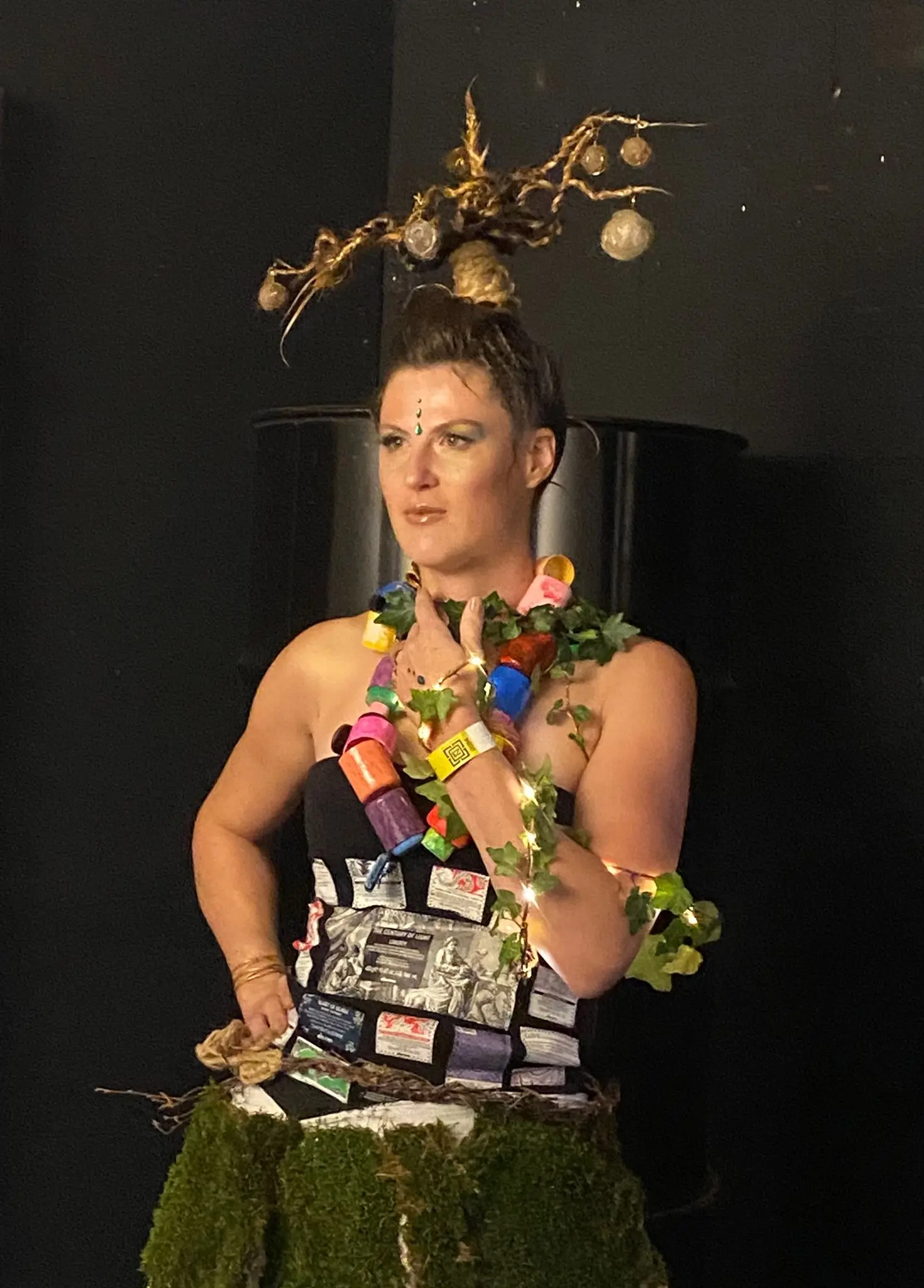 Donna con un abito creativo realizzato con materiali riciclati, adornata da rami e luci, in piedi davanti a uno sfondo scuro. Ha un'espressione seria e indossa gioielli colorati.