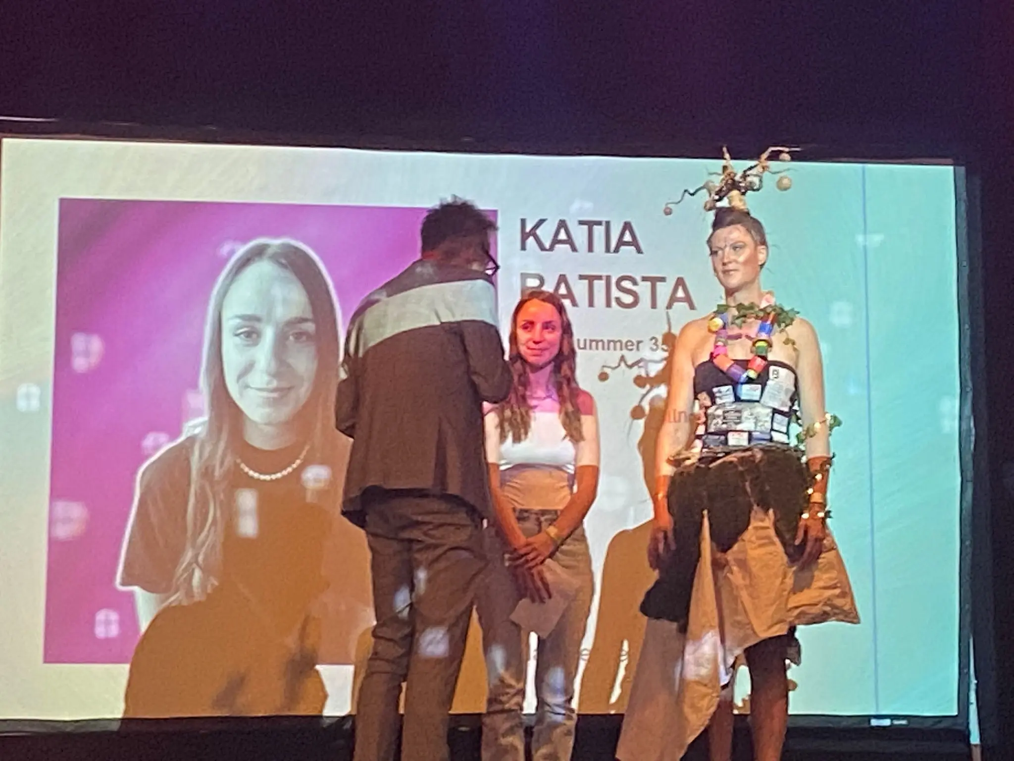 Una donna in abbigliamento artistico e creativo si trova sul palco, circondata da due persone. Dietro di lei c'è uno schermo con il nome "Katia Batista" e un'immagine di una giovane donna. L'atmosfera è vivace e colorata.