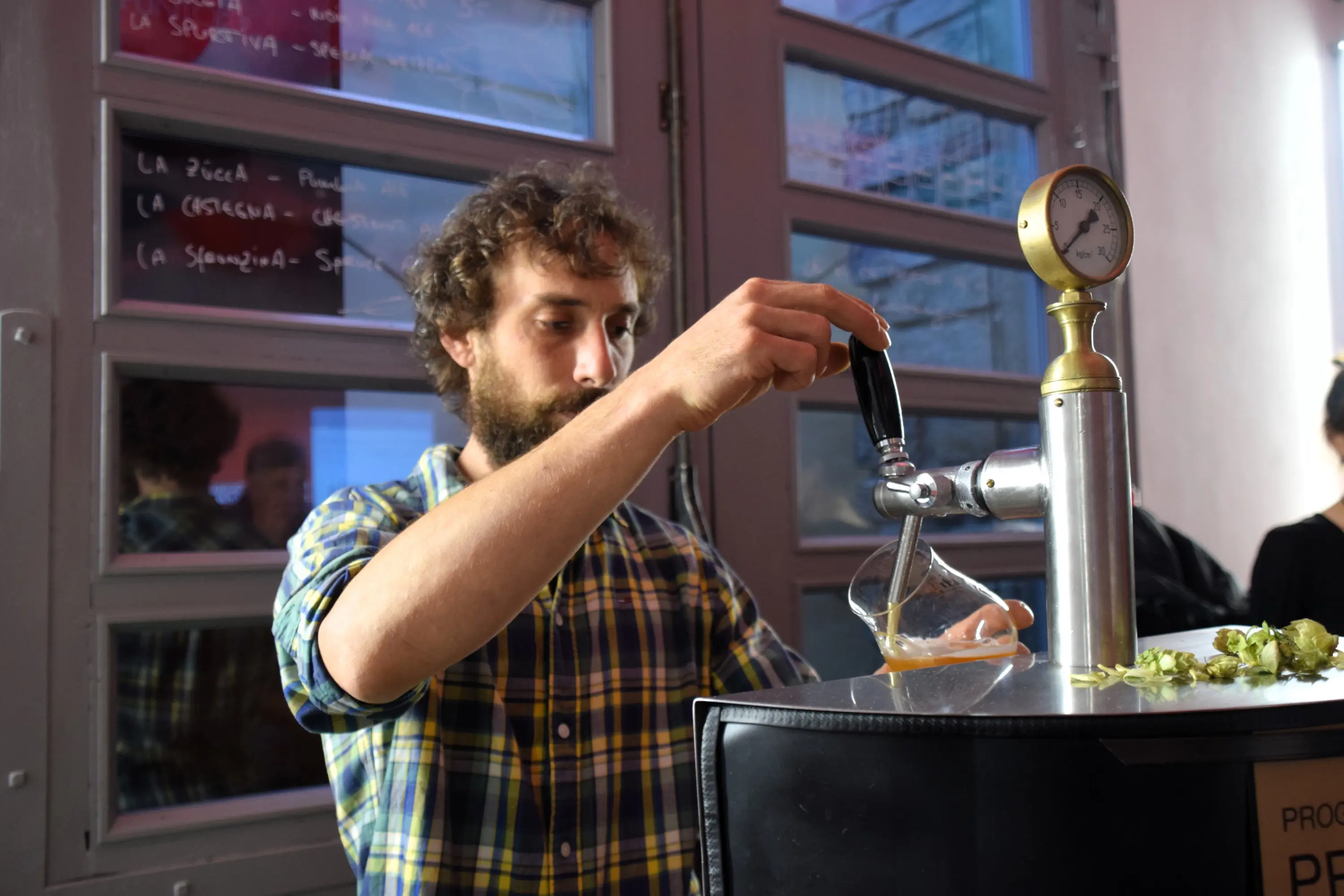 Un uomo con una barba folta e una camicia a quadri versa una bevanda da un rubinetto in un ambiente modernista. Sullo sfondo si vedono scritte su una lavagna.