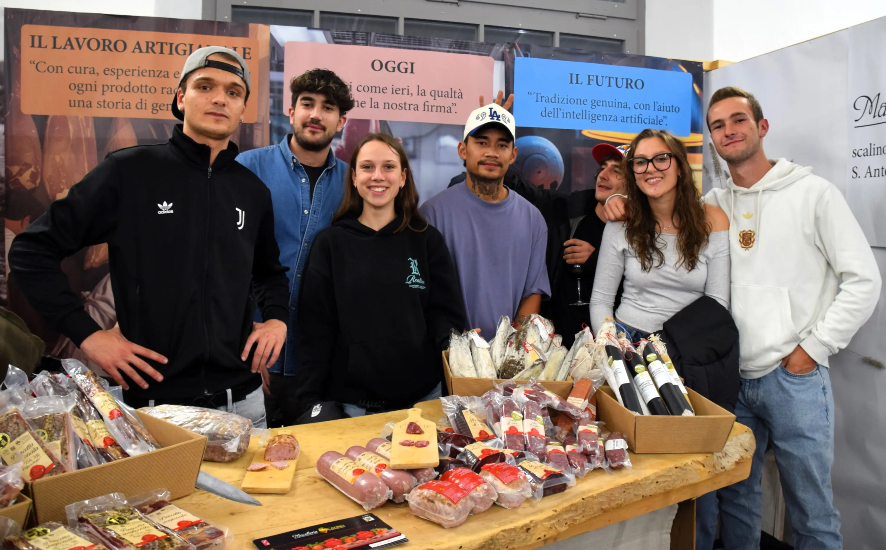 Gruppo di sei persone sorridenti in un evento, posando davanti a un tavolo pieno di salumi e formaggi. Sullo sfondo, pannelli informativi sul lavoro artigianale e il futuro della tradizione alimentare.