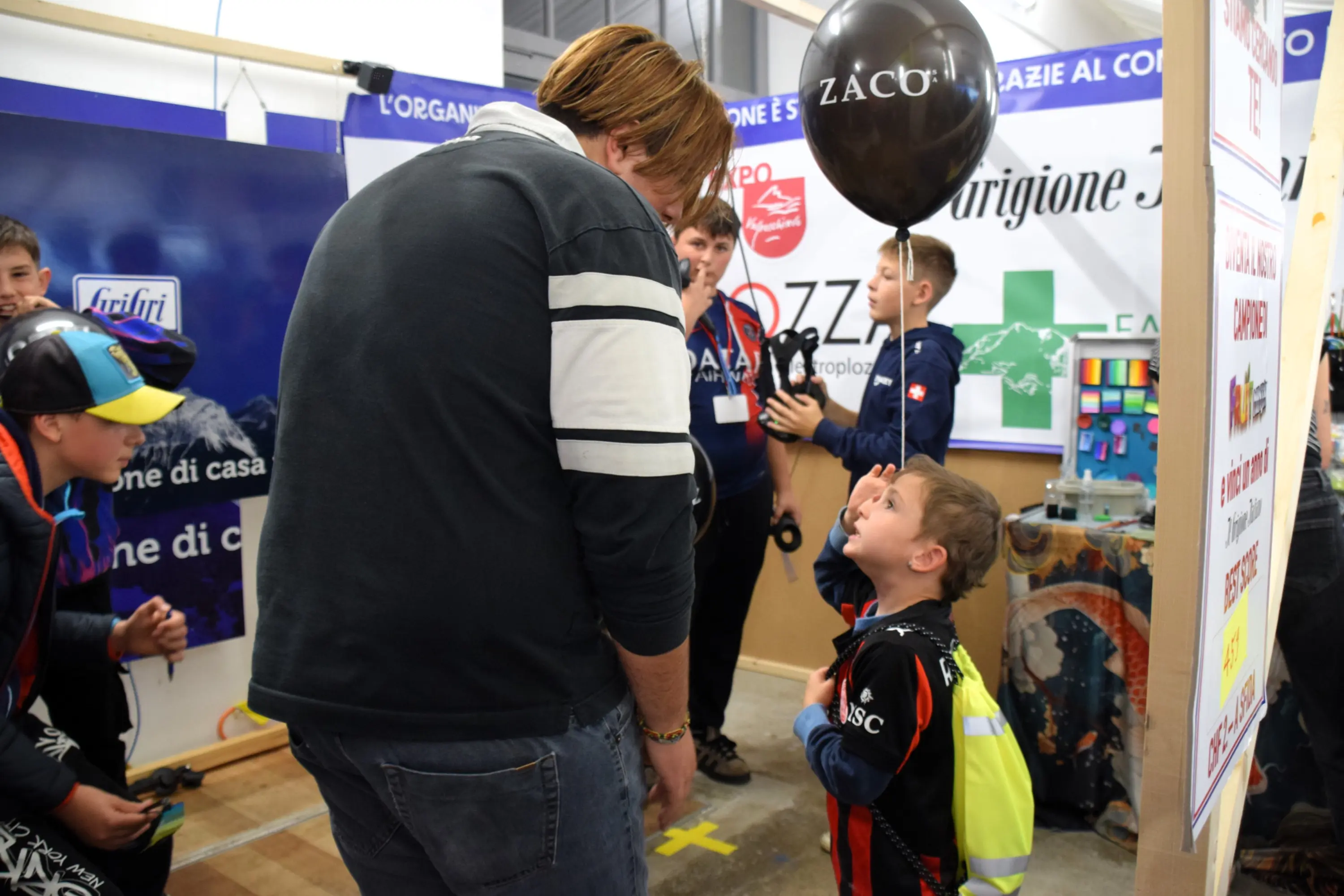 Un bambino con un palloncino nero guarda un adulto mentre sorride. Sono entrambi all'interno di un evento, con vari stand e oggetti esposti sullo sfondo.
