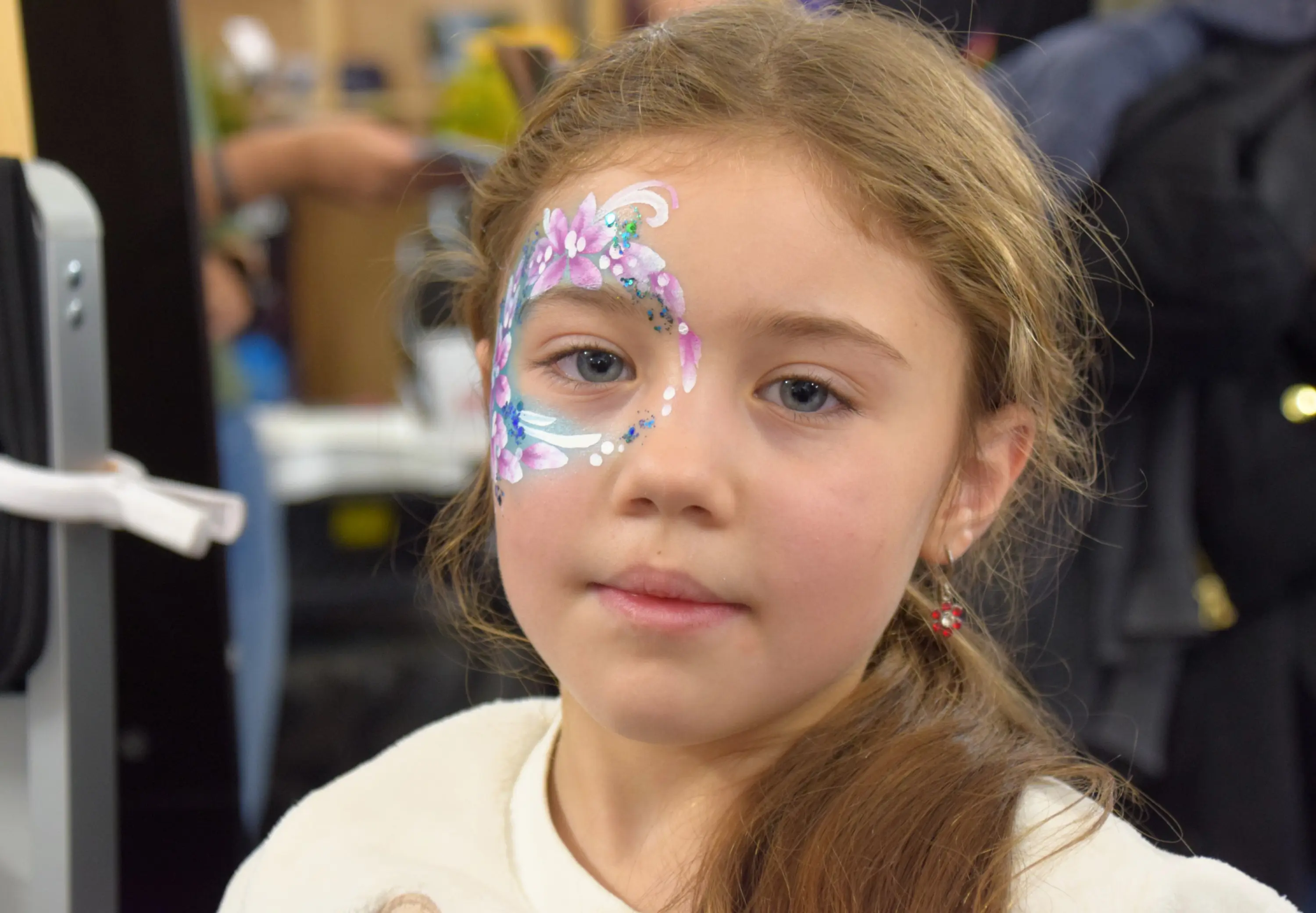 Bambina con viso dipinto, decorato con fiori e glitter colorati. Ha capelli lunghi e castani e indossa una maglietta chiara.