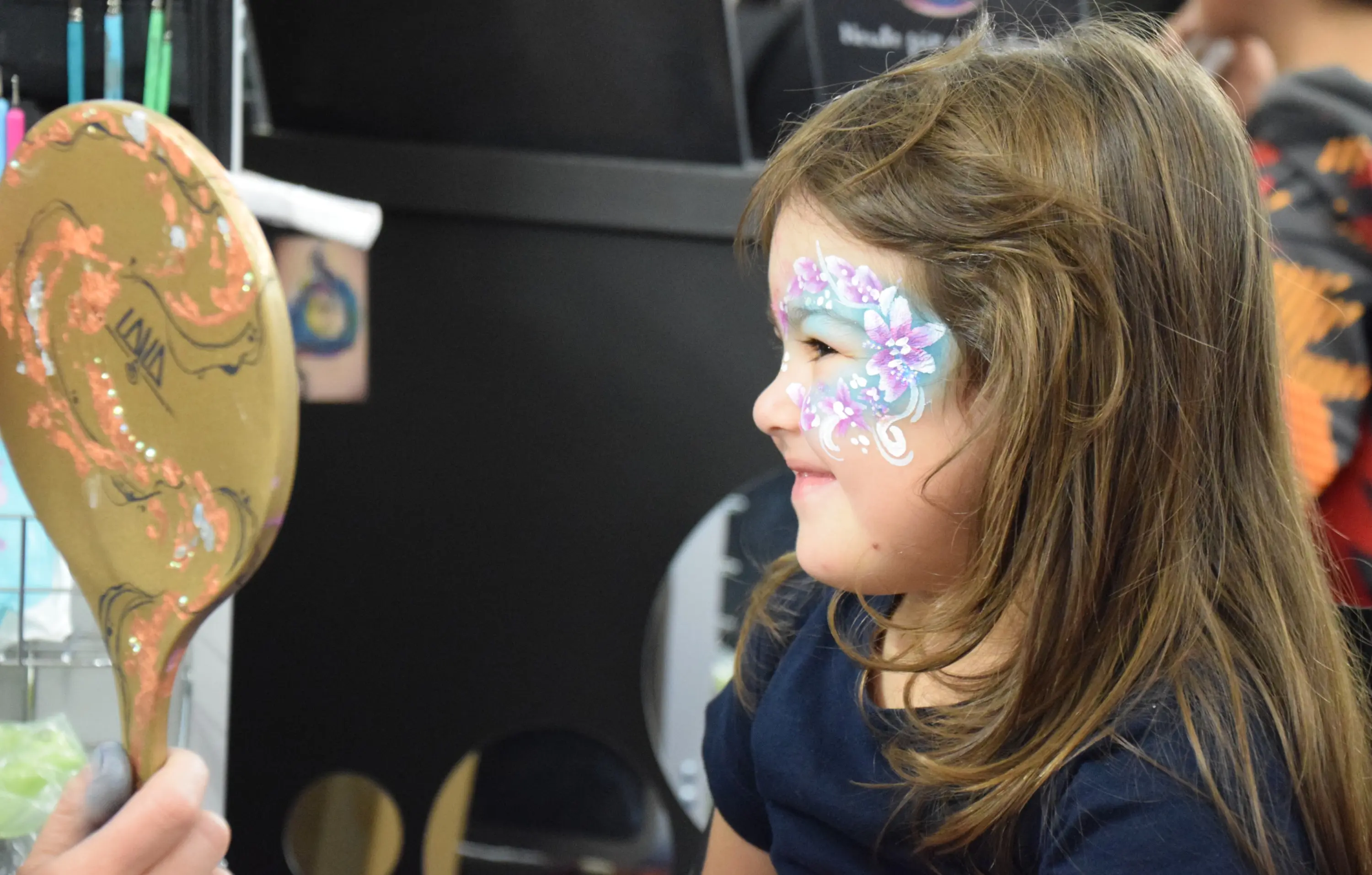 Una bambina sorridente osserva il suo riflesso in uno specchio, con il viso dipinto da un artista. Il trucco include fiori colorati e decorazioni intorno agli occhi.