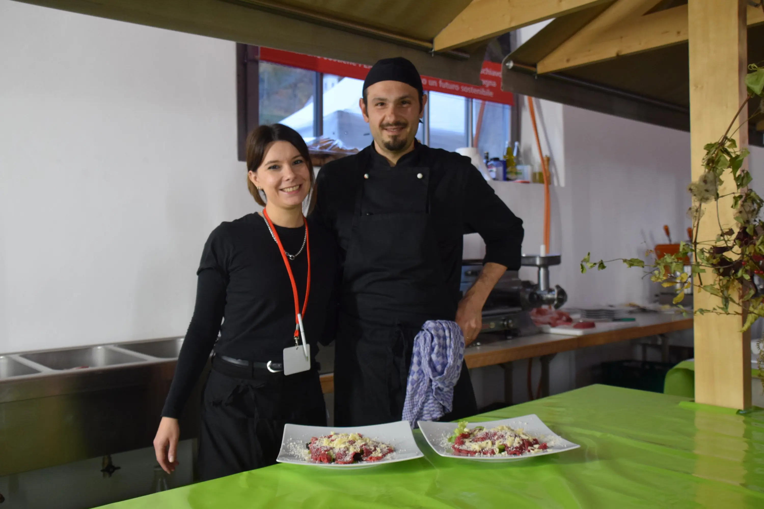 Due persone sorridenti in un ambiente di cucina, indossano abbigliamento da cuoco. Sono accanto a due piatti di cibo ben presentati su un tavolo verde.