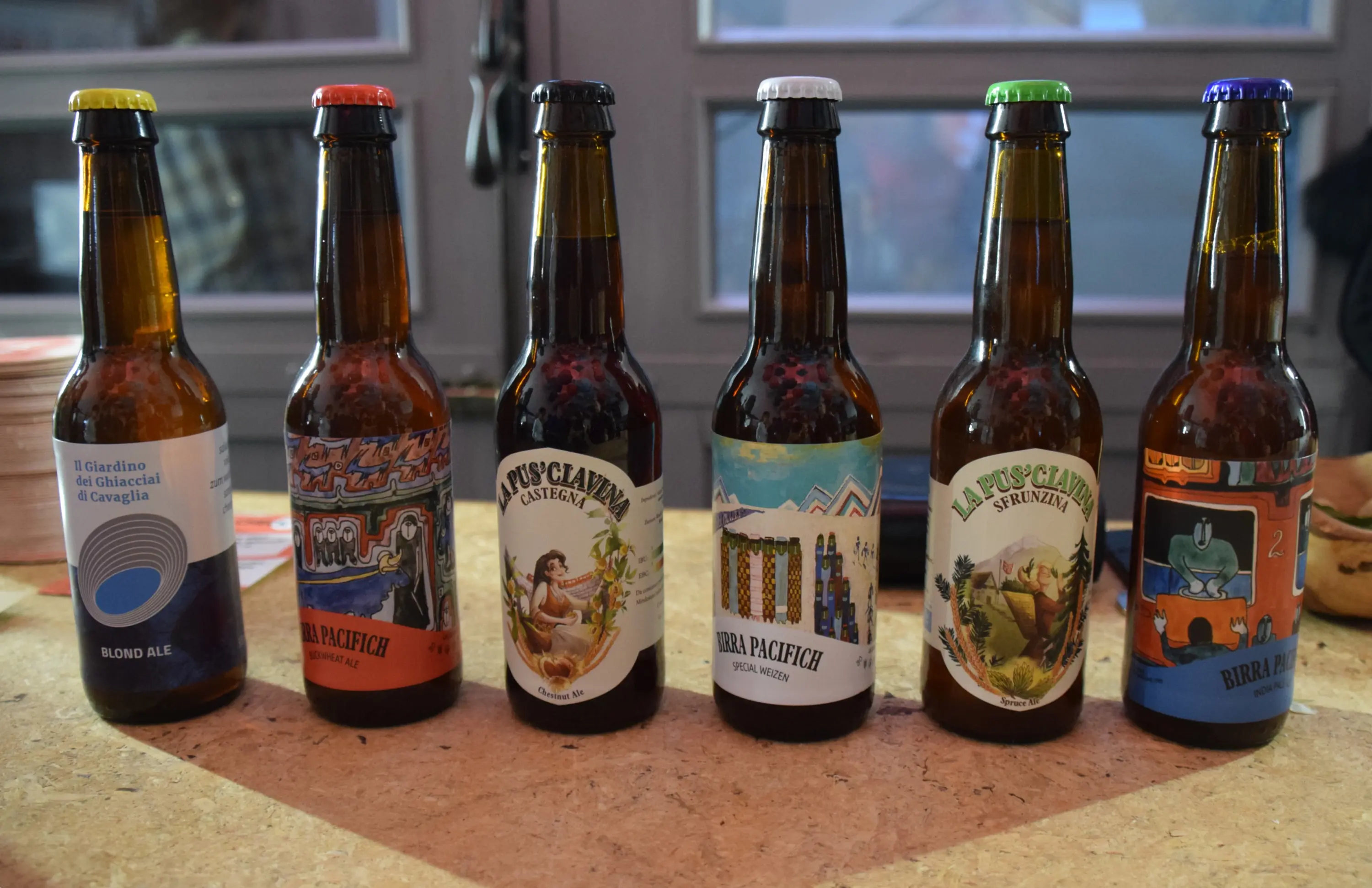 Sette bottiglie di birra allineate su un tavolo. Le etichette mostrano vari design colorati, inclusi paesaggi e figure stilizzate. Ogni bottiglia ha un colore di tappo diverso.