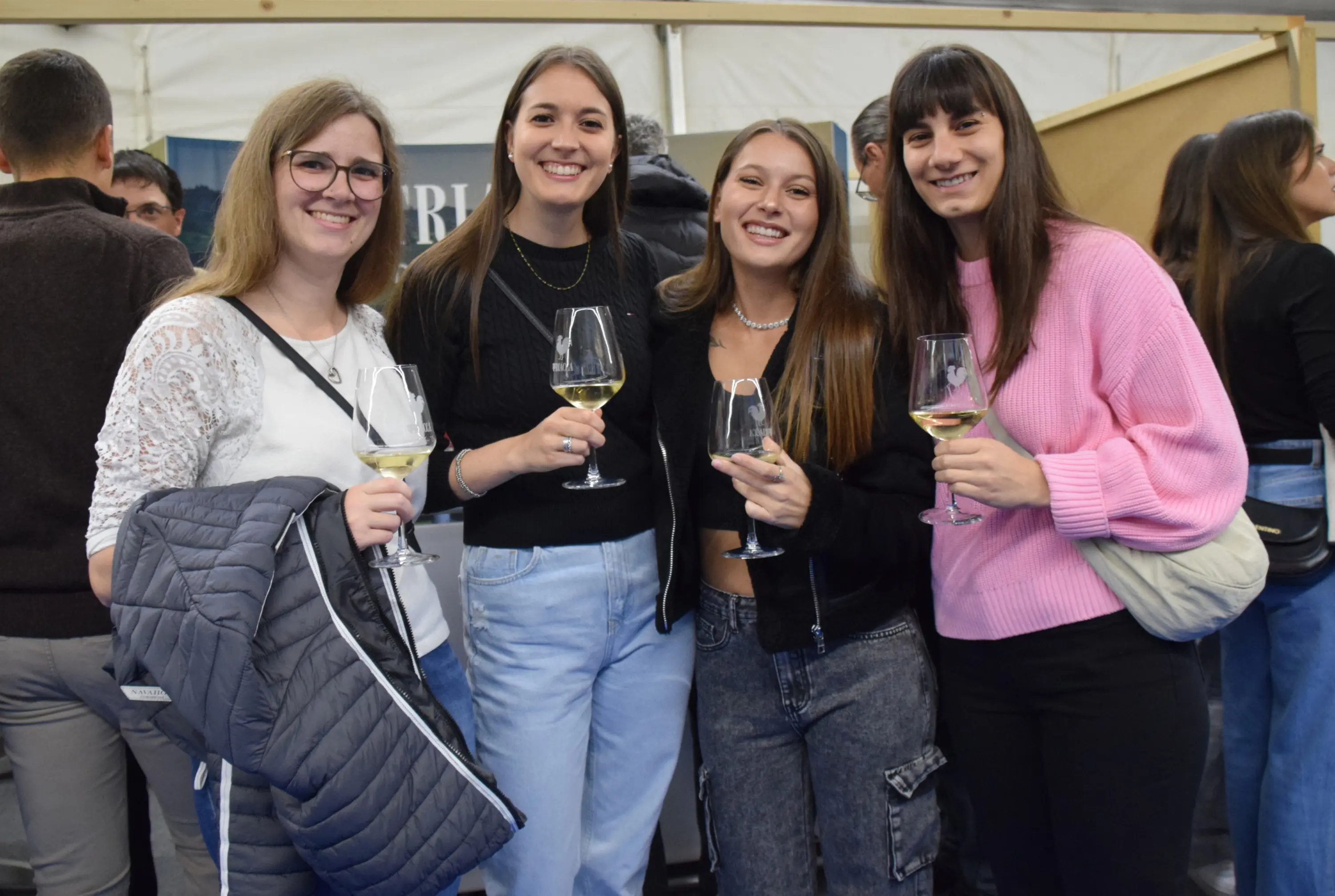 Quattro giovani donne sorridenti, ciascuna con un bicchiere di vino in mano, si trovano in un ambiente festoso. Indossano abiti casual e si divertono in compagnia.