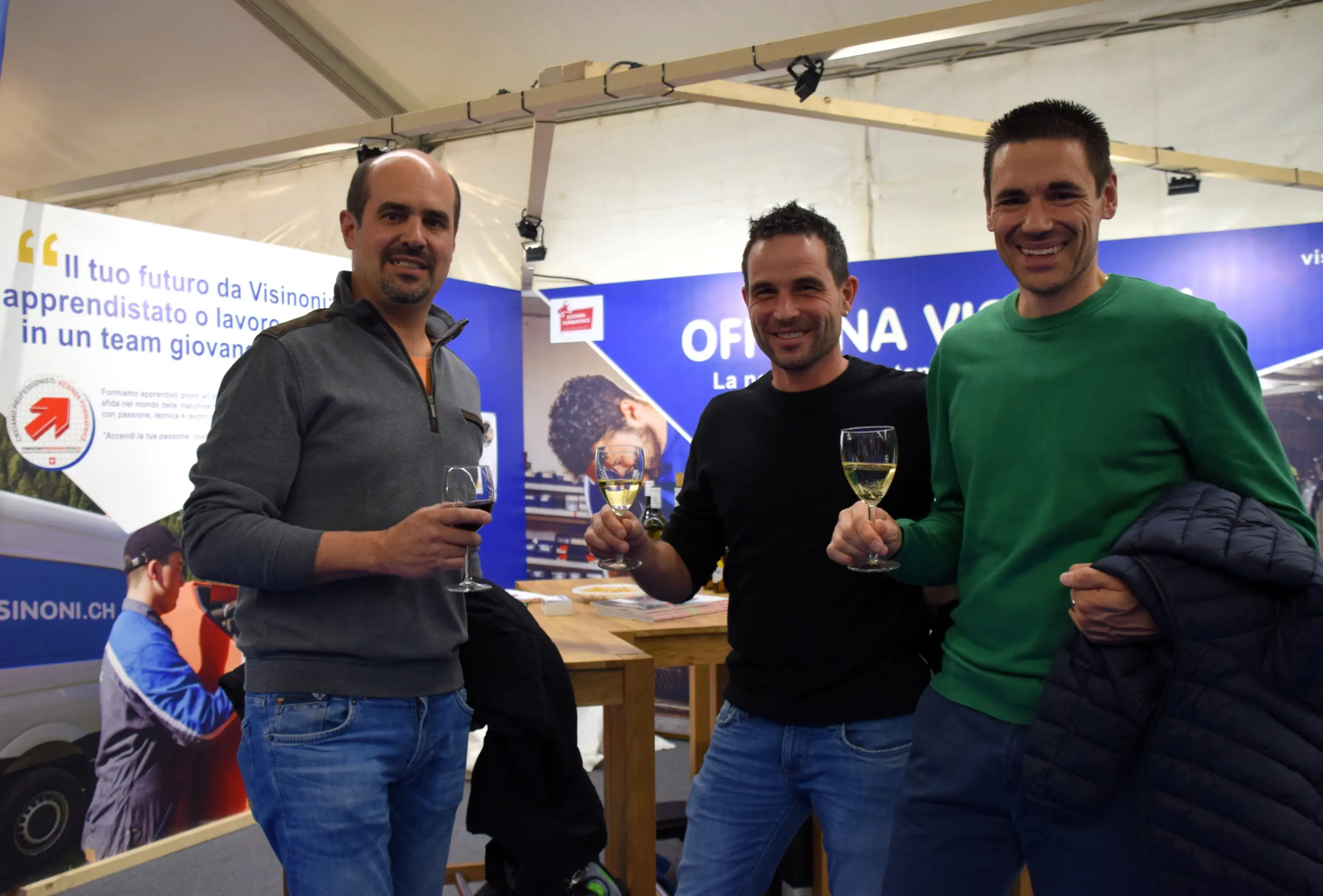 Tre uomini sorridenti tengono in mano dei calici di vino, in un ambiente fieristico. Sullo sfondo si intravedono banner con scritte e informazioni.