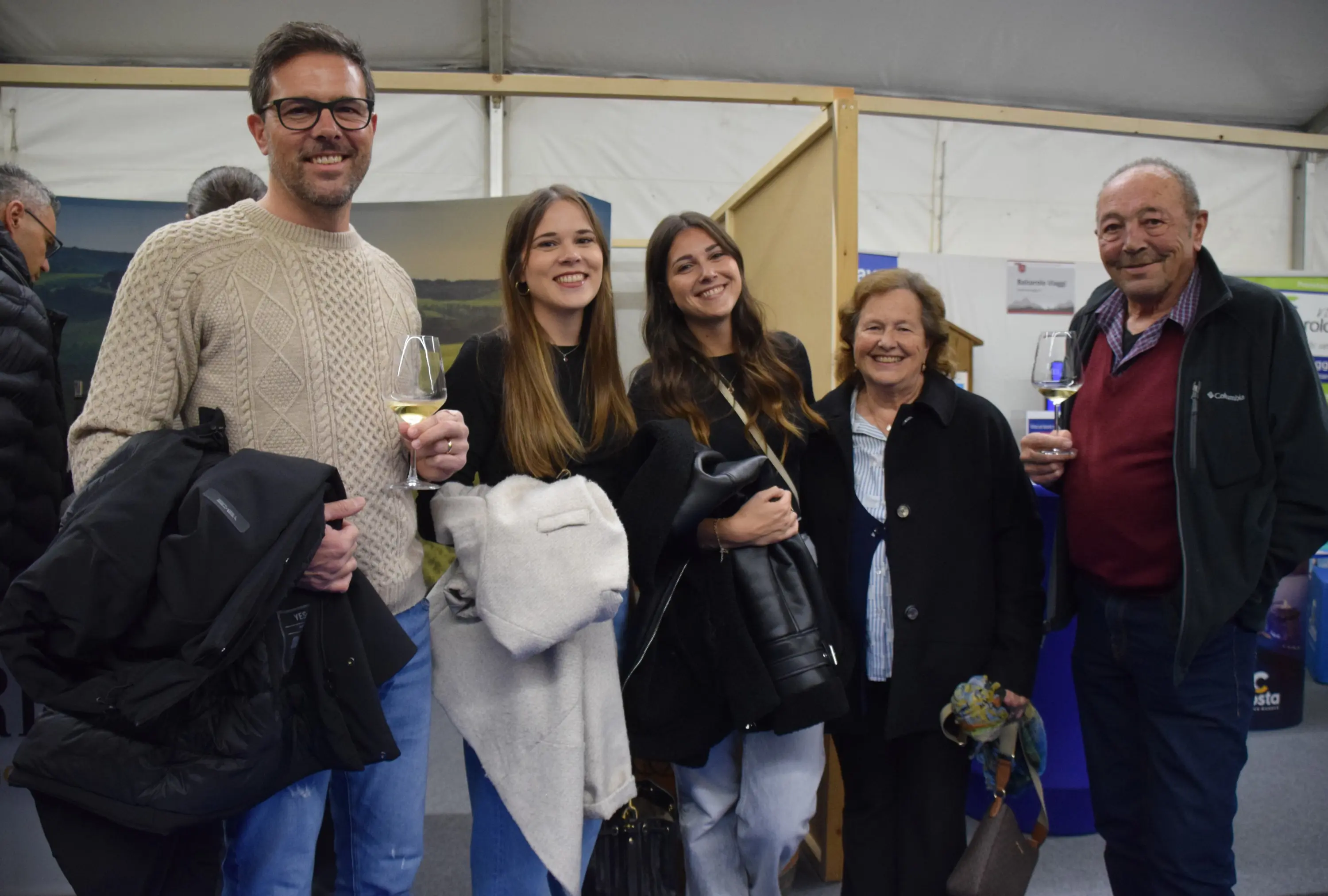 Un gruppo di cinque persone sorride, tenendo in mano dei bicchieri di vino. Sono in un ambiente festoso, probabilmente a un evento, e sfoggiano abiti casual.