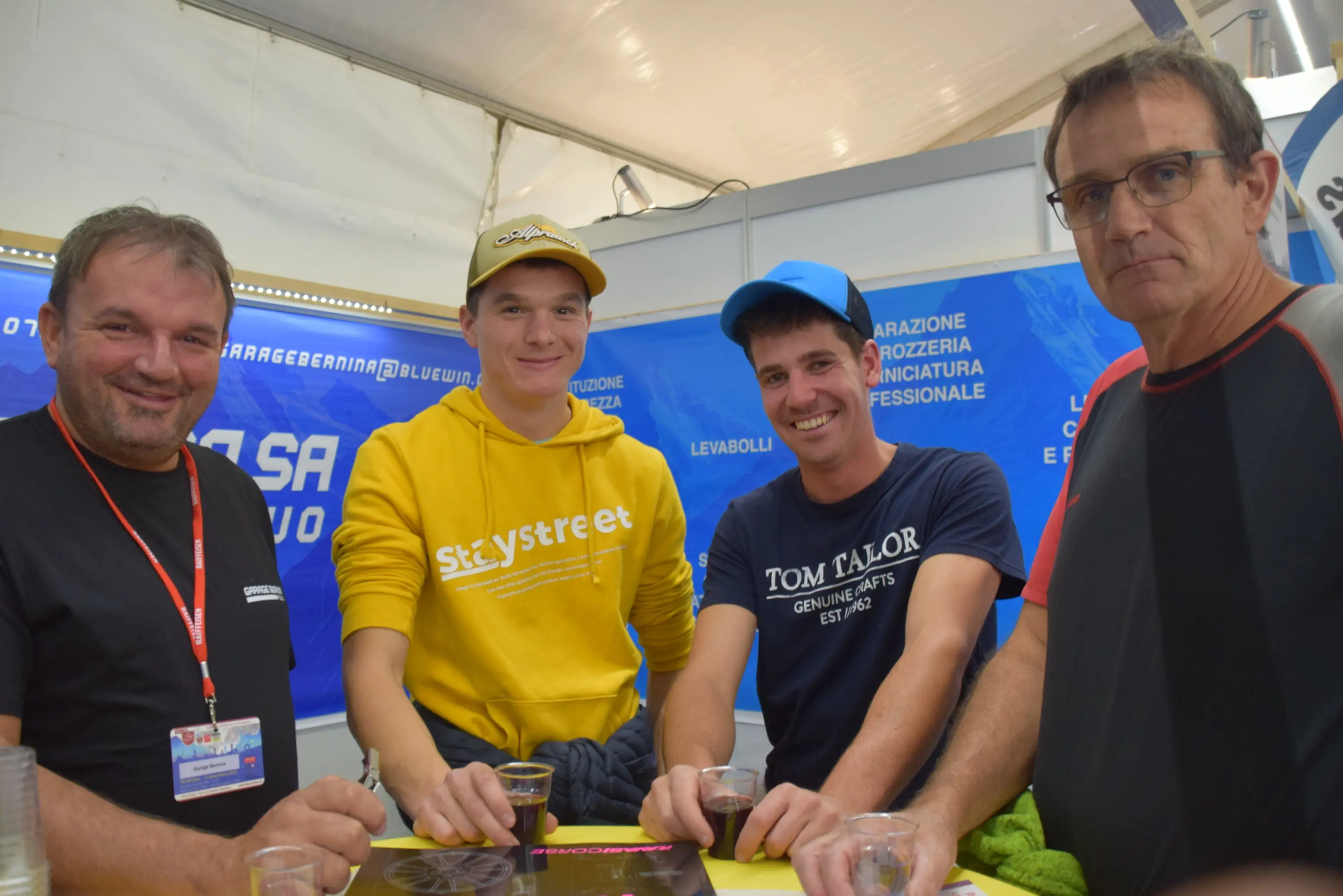 Quattro uomini sorridenti si radunano attorno a un tavolo, con bicchieri in mano. Indossano abbigliamento casual, con alcuni berretti. Nel retro, si intravedono striscioni e pubblicità.