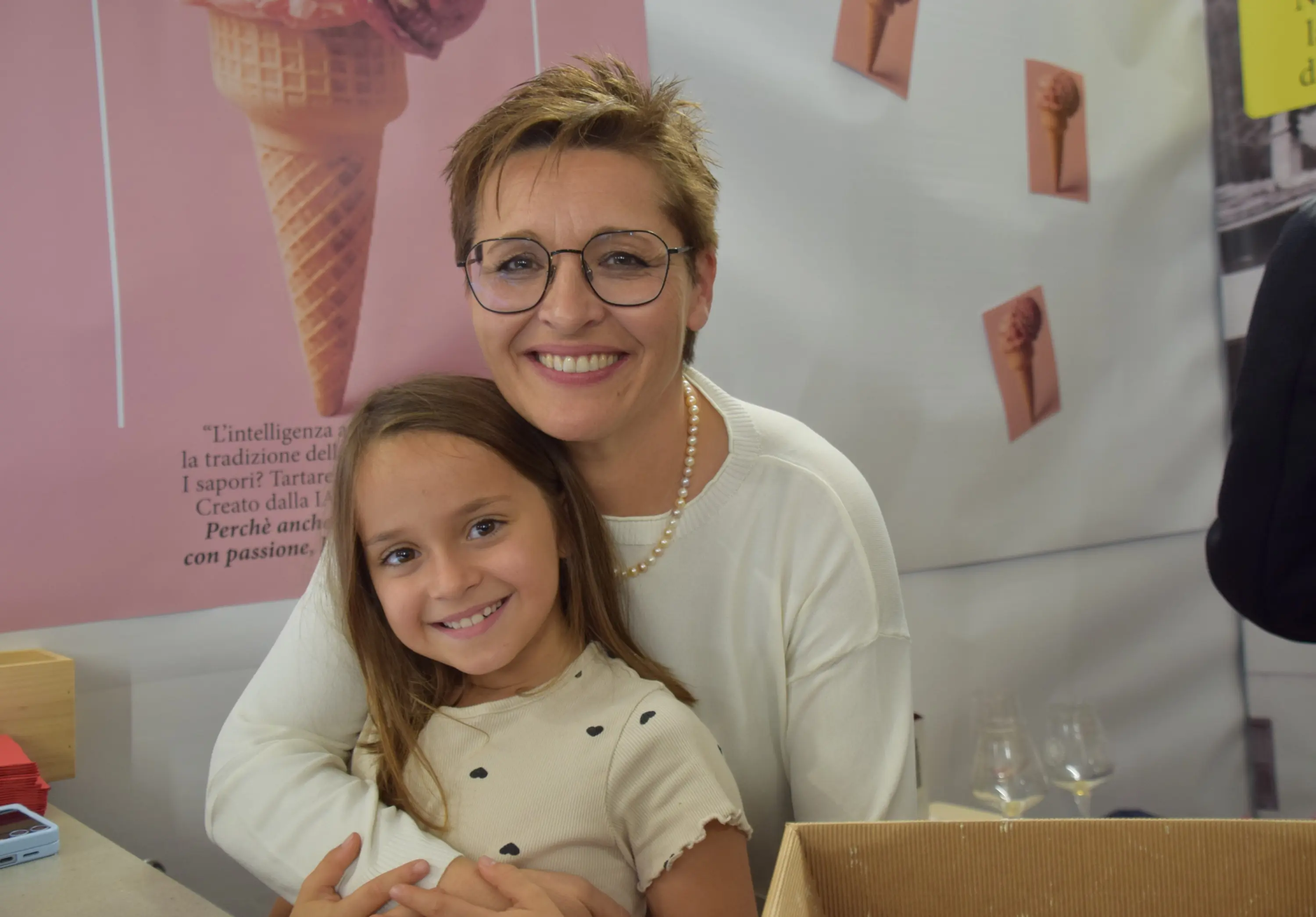Una donna sorridente con occhiali abbraccia una bambina in un evento. Sullo sfondo, un poster con coni gelato. Entrambe sembrano felici e stanno godendo il momento insieme.
