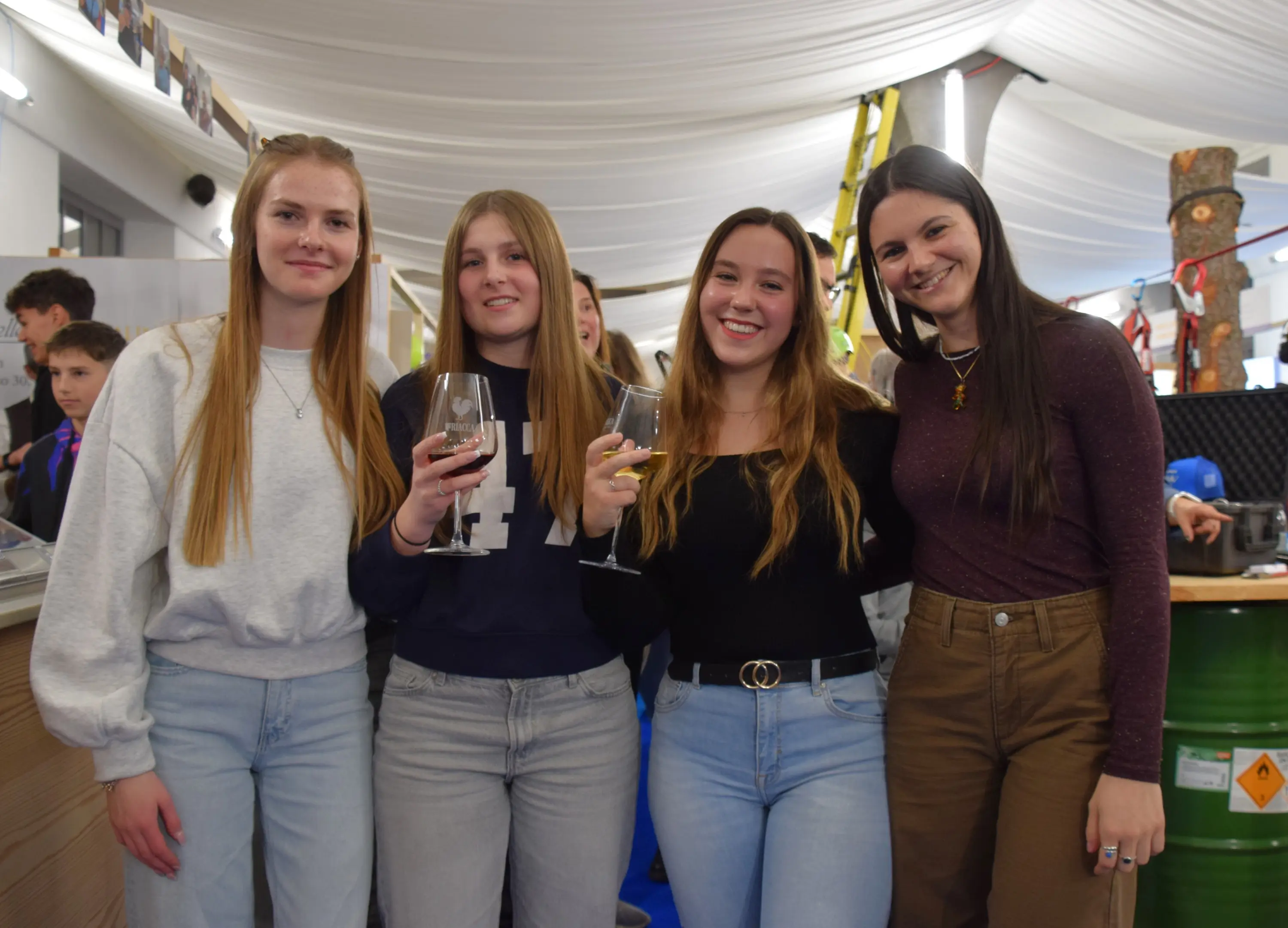 Quattro ragazze sorridenti si trovano in un ambiente festivo, alcune con bicchieri in mano. Sono vestite casualmente; due indossano maglioni e due top a maniche lunghe. Sullo sfondo si intravede un gruppo di persone e decorazioni