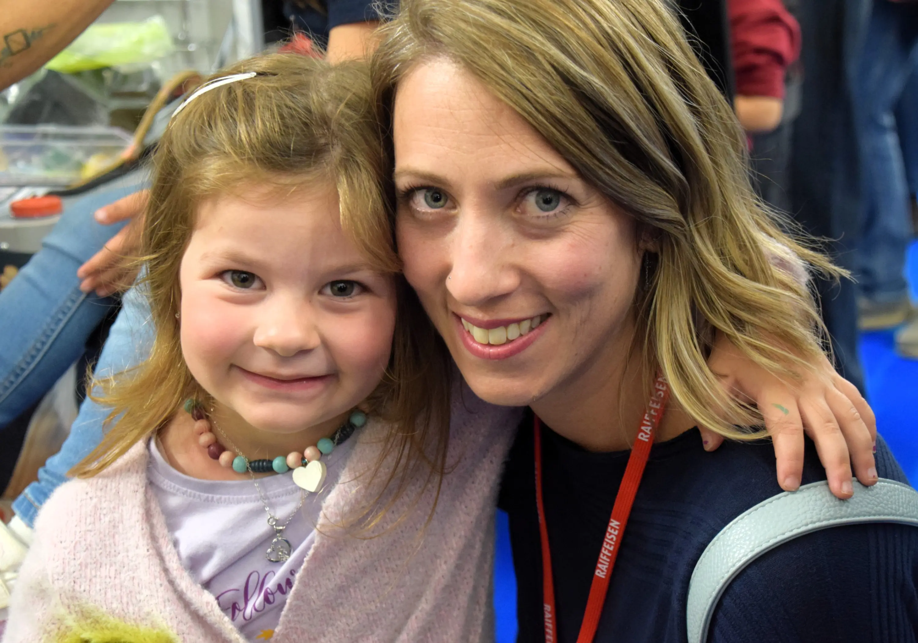 Una donna sorride accanto a una bambina. La donna ha i capelli biondi ondulati e indossa una maglietta blu con un pass per un evento. La bambina, con i capelli castani, è abbracciata a lei e indossa una maglia