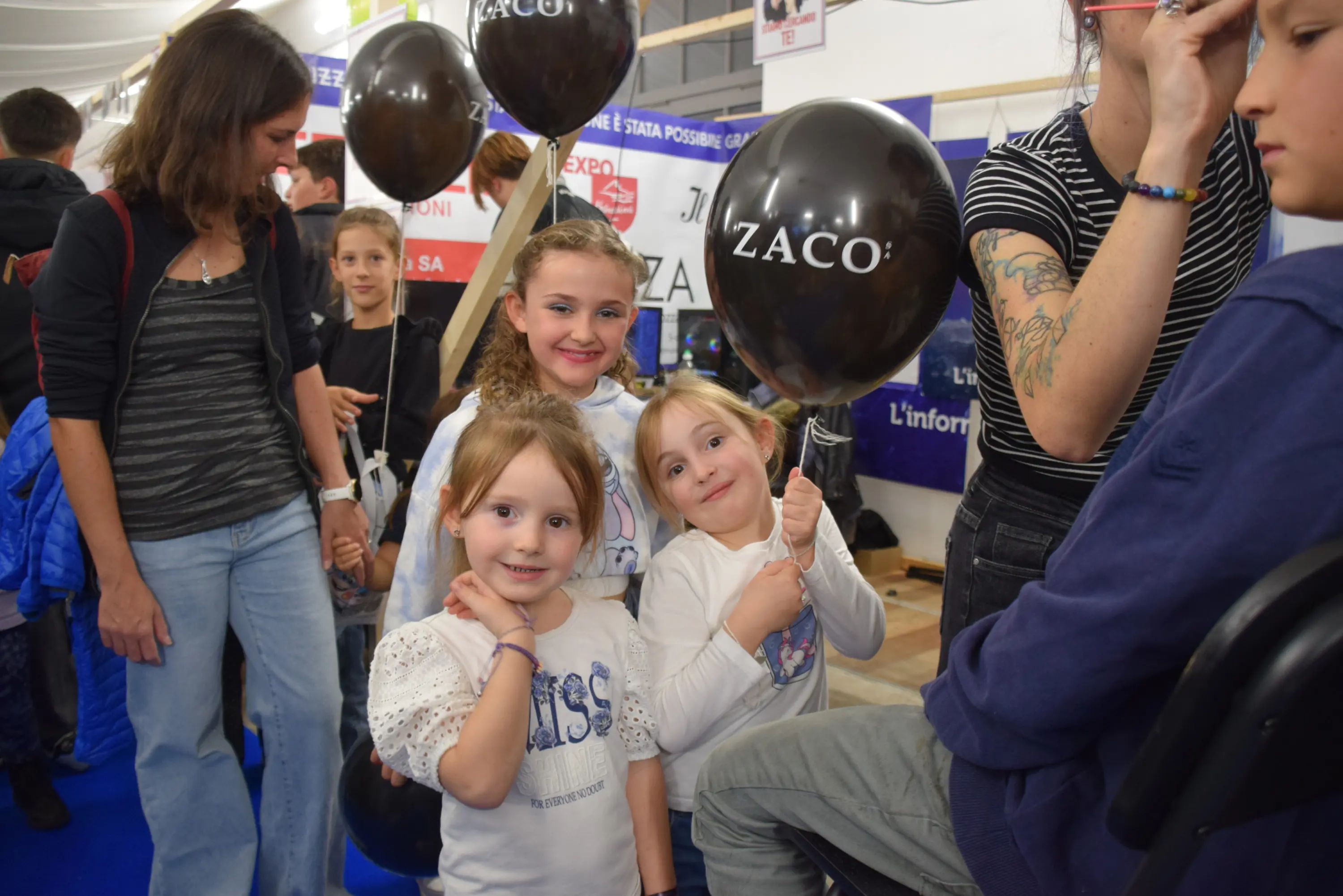 Tre bambine sorridenti tengono in mano palloncini neri, mentre altre persone si trovano sullo sfondo. L’atmosfera è festosa e colorata.