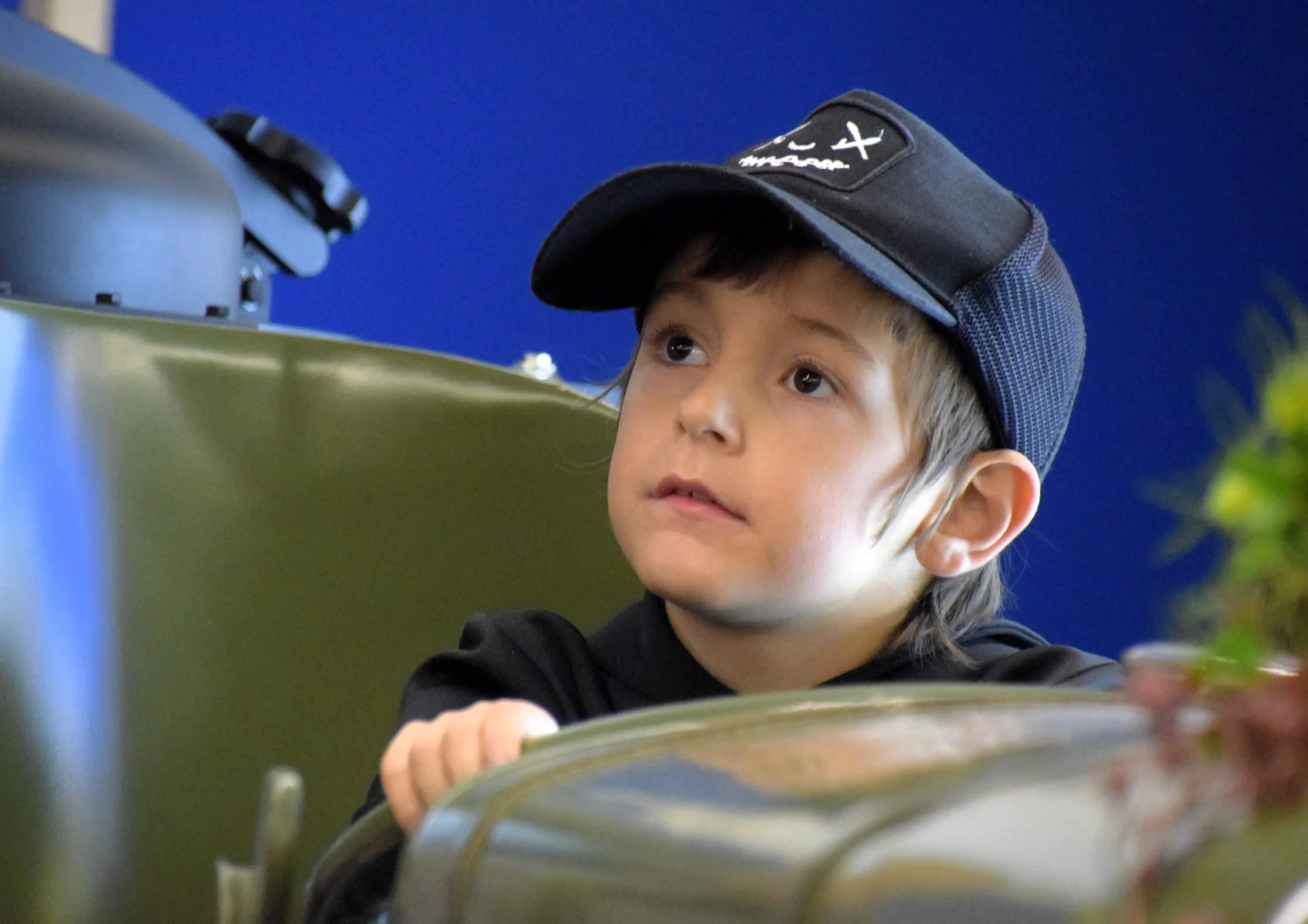 Un bambino con un cappellino scuro guarda attentamente. È seduto in un veicolo verde, con un'espressione curiosa sul volto. Sullo sfondo si intravede una parete blu.