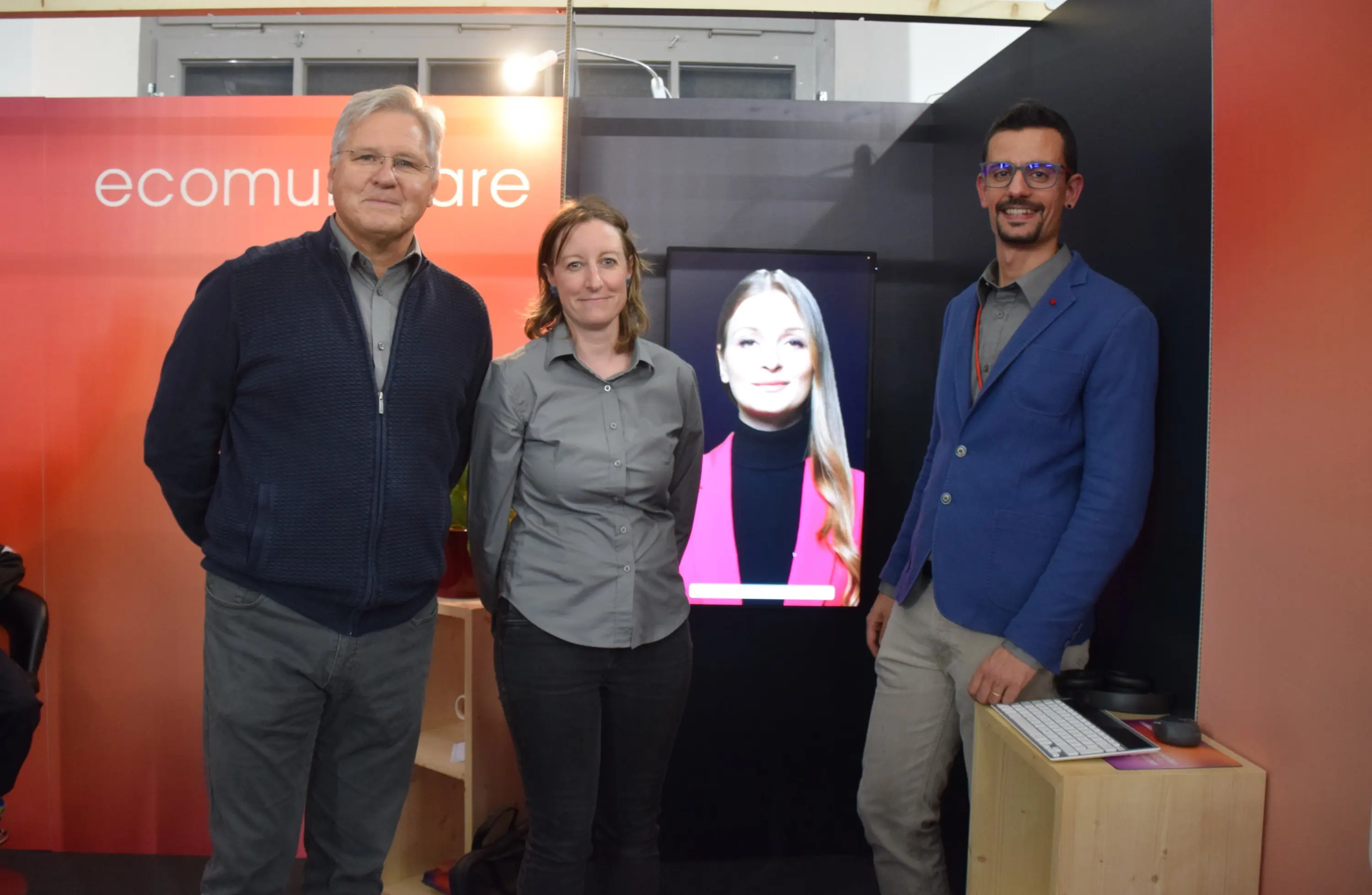 Tre persone sorridenti si trovano davanti a uno schermo con un'immagine di una donna. Due di loro indossano camicie grigie e l'altro un abito blu. Dietro c'è uno sfondo rosso con la scritta "ecomunicare".