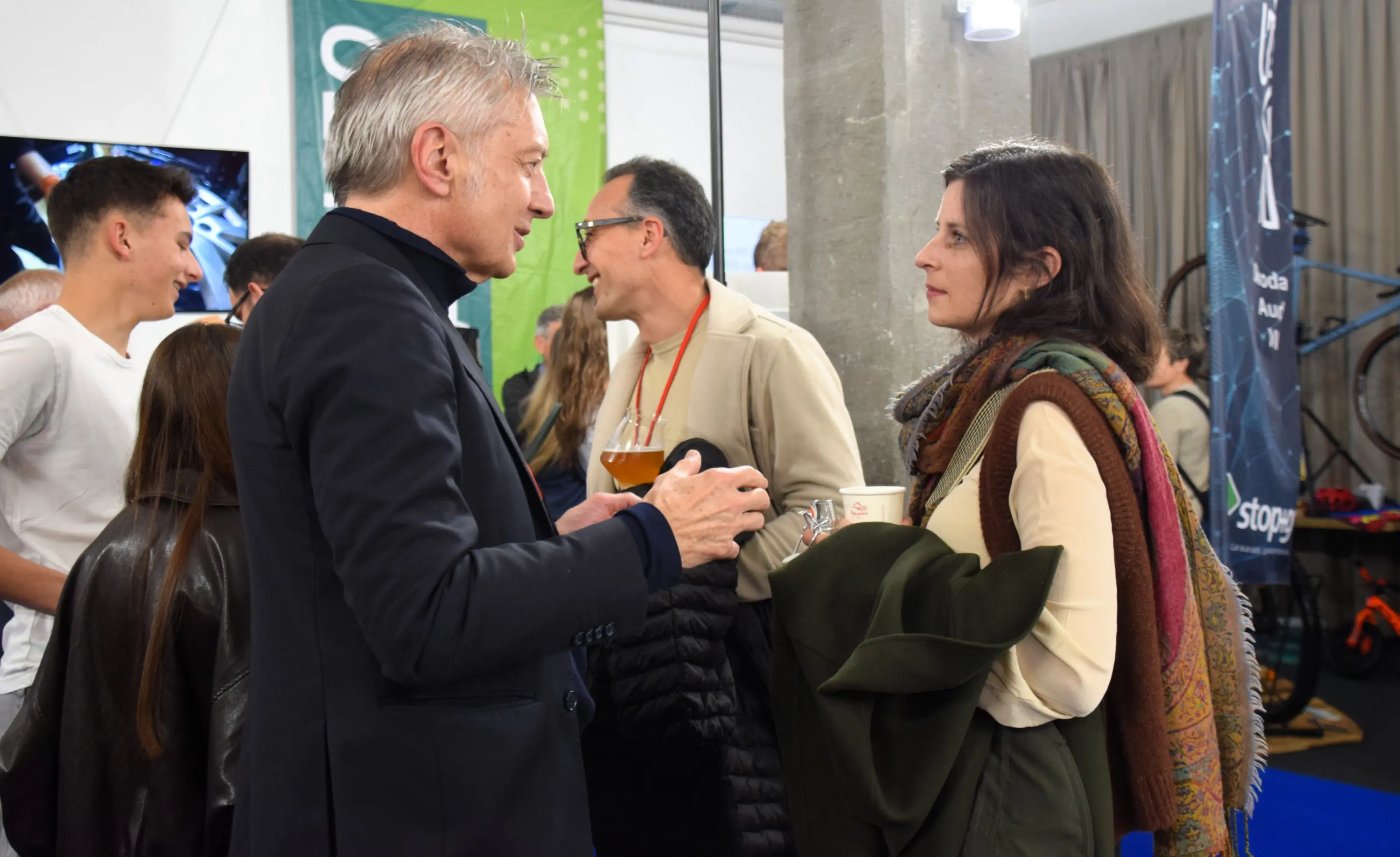 Due persone conversano in un ambiente affollato. L'uomo, con capelli grigi, indossa un abito scuro e tiene un bicchiere in mano. La donna, con capelli scuri e un foulard colorato, ascolta attentamente. Sullo sfondo,