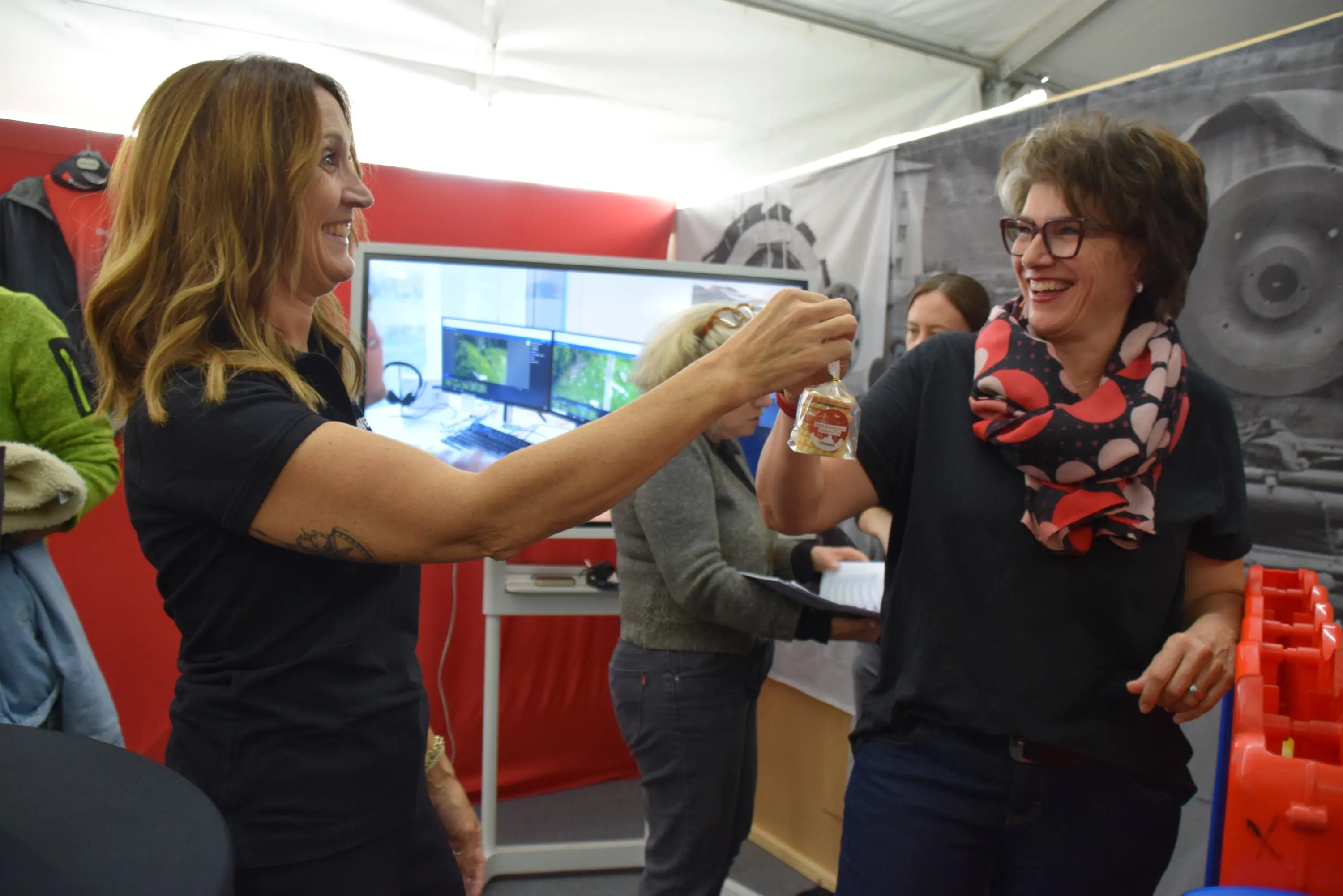 Due donne sorseggiano un drink, sorridendo e brindando tra di loro. Sullo sfondo si notano alcune persone e un monitor con immagini.