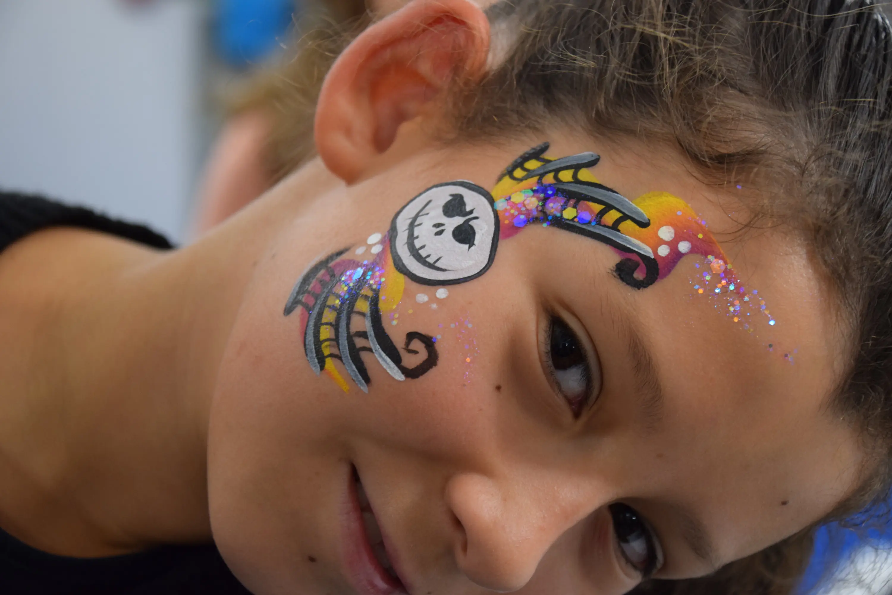 Una bambina sorride con un volto dipinto, mostrando un disegno di un teschio e decorazioni colorate e glitterate.