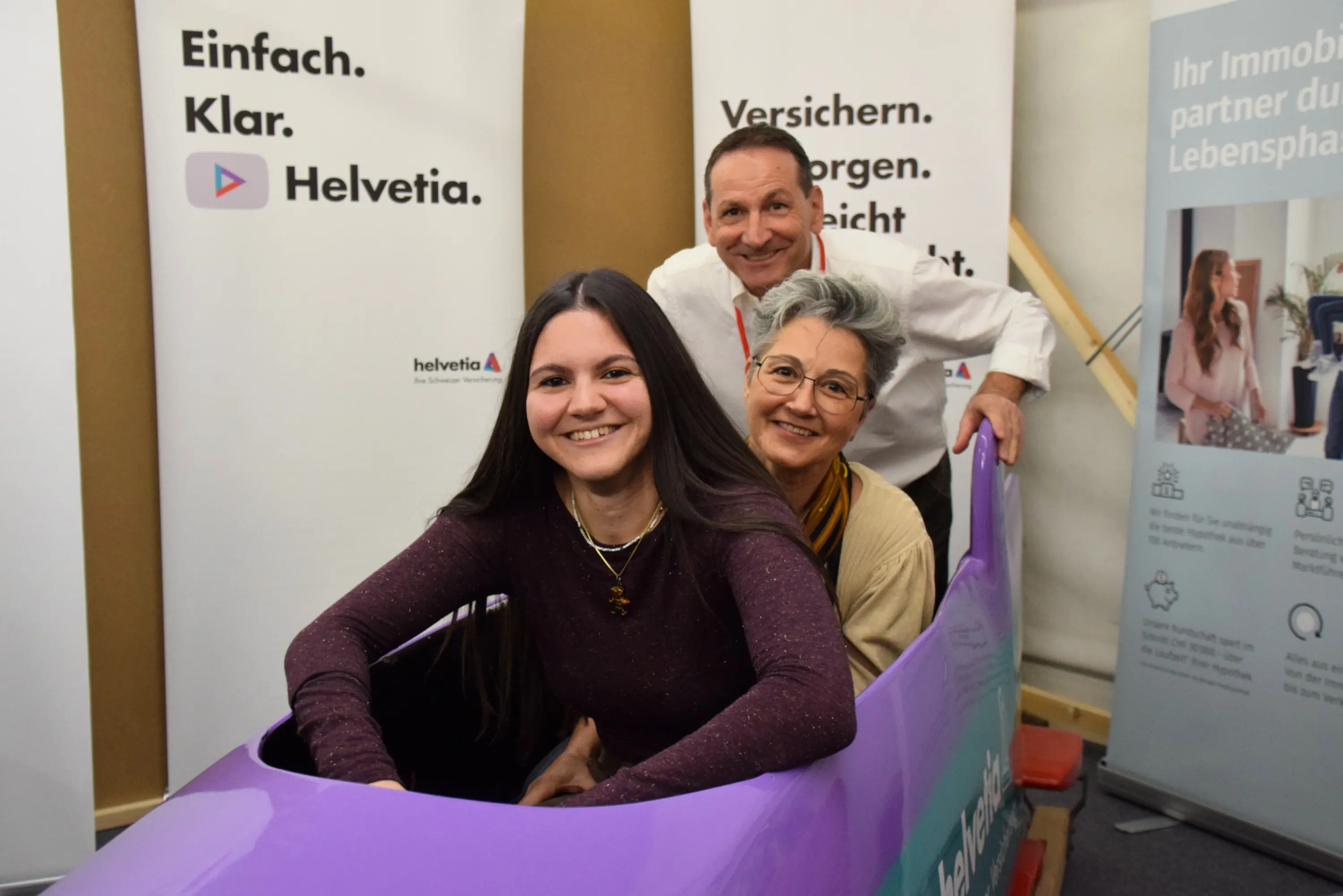 Tre persone sorridenti sono sedute in un simulatore a forma di auto, circondate da poster promozionali di Helvetia. Due persone sono sedute nella parte anteriore e una nella parte posteriore, tutte si divertono.