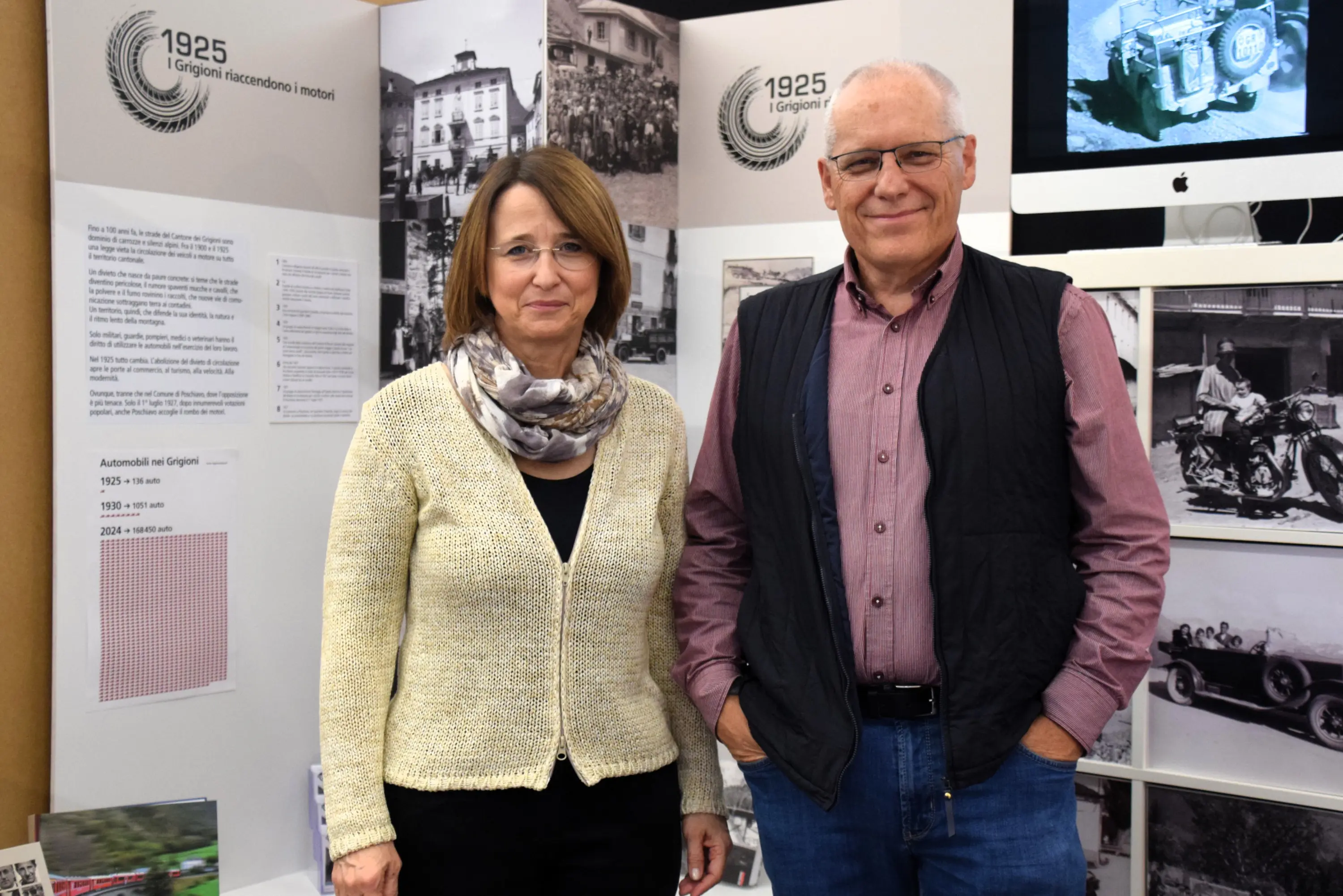 Due persone sorridenti, una donna con sciarpa e maglione chiaro e un uomo con occhiali e gilet scuro, posano davanti a una mostra con pannelli informativi su storie automobilistiche.