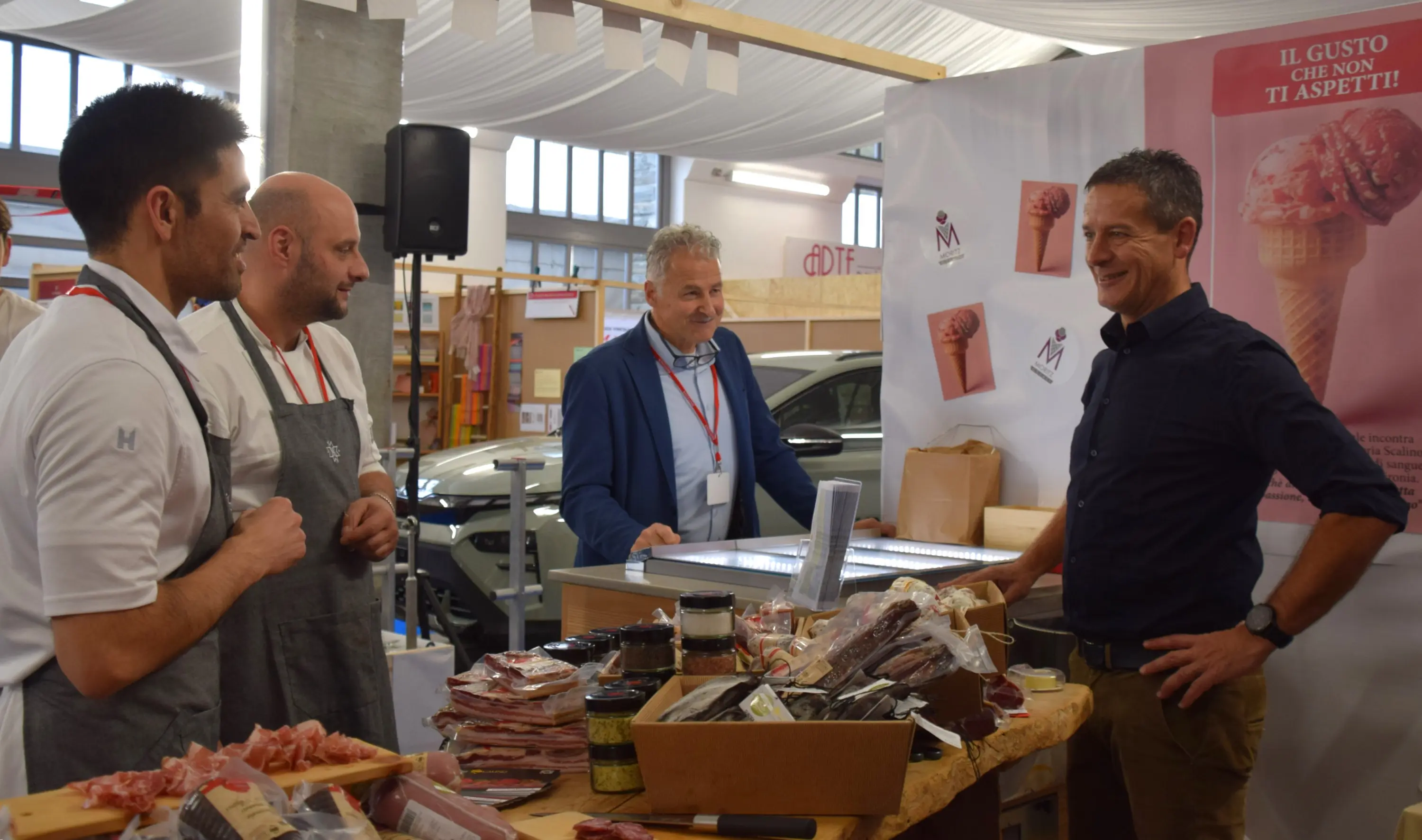 Un gruppo di uomini si confronta a uno stand fieristico con prodotti gastronomici. Due uomini indossano grembiuli e parlano con un altro uomo, mentre un quarto osserva. Sullo sfondo c'è un cartellone con un messaggio promozionale.