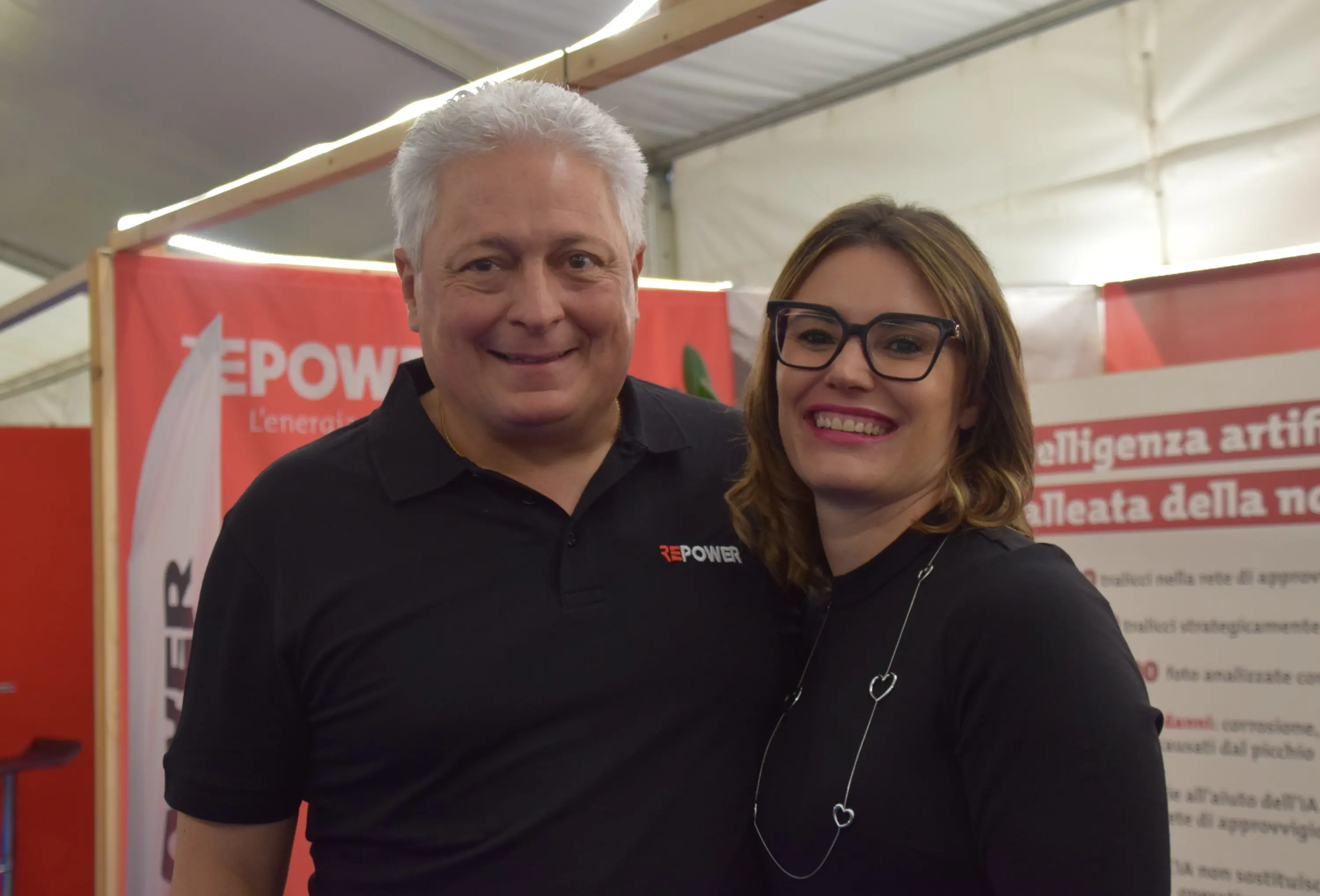 Due persone sorridono, una uomo con una polo nera con il logo "EPower" e una donna con occhiali e una maglia nera. Sullo sfondo si intravedono elementi di un evento o stand.